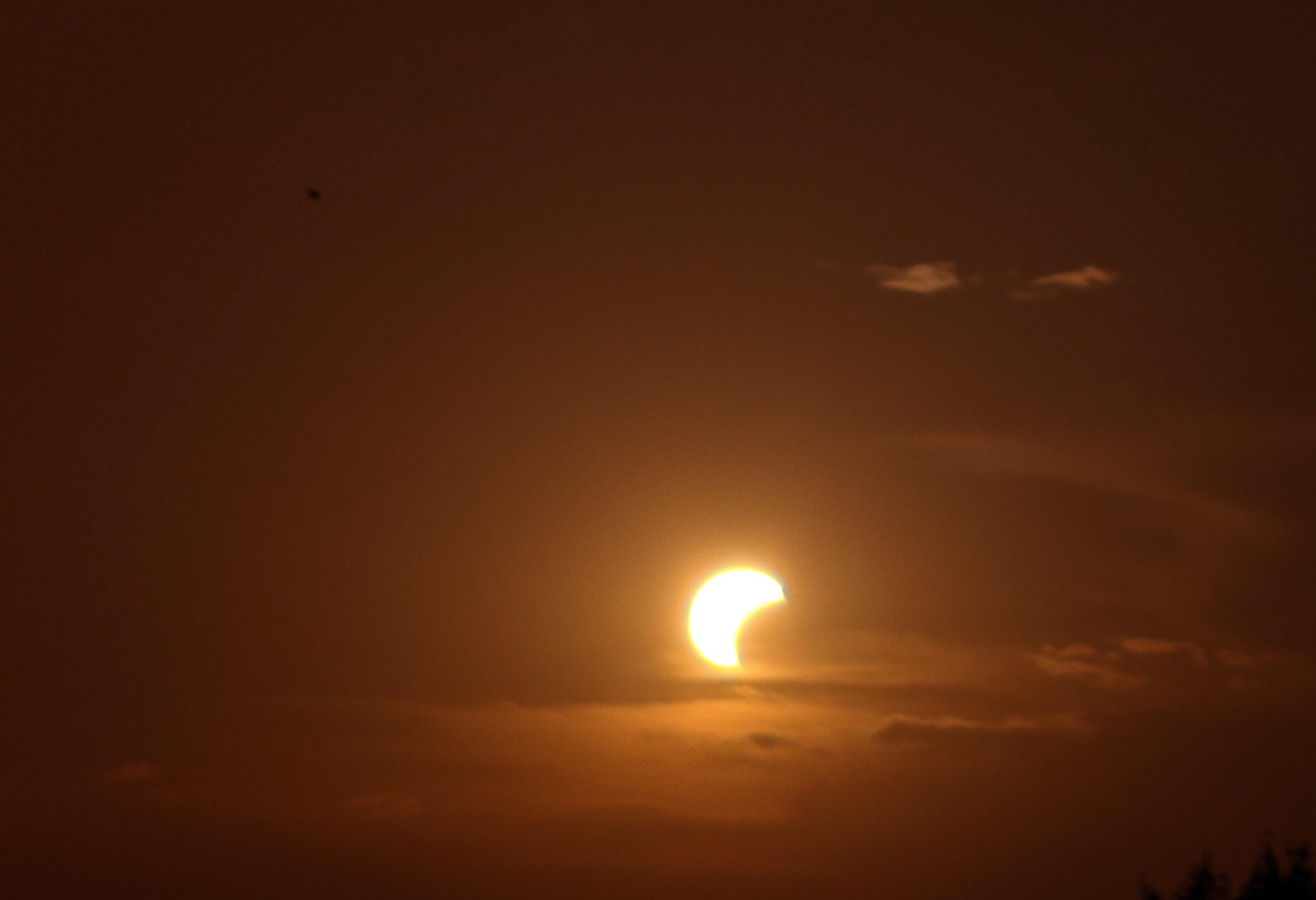 L'immagine mostra un suggestivo tramonto durante un'eclissi parziale di sole, caratterizzato da un'atmosfera calda e drammatica.Ecco i dettagli principali della scena:Il Sole ed l'EclissiForma a falce: Il sole non è un disco pieno, ma appare ridotto a una lucente falce dorata rivolta verso sinistra.Copertura: La luna copre gran parte della stella, lasciando visibile solo una sezione curva molto luminosa.Il Cielo e i ColoriTonalità calde: L'intero cielo è dominato da sfumature che vanno dall'arancione scuro al marrone bruciato, tipiche del crepuscolo o di un'atmosfera densa di foschia.Nuvole basse: Sottili strati di nuvole scure tagliano orizzontalmente la base del sole, parzialmente illuminate dal basso.Nuvole alte: Piccoli filamenti di nuvole più chiare galleggiano in alto a destra, catturando gli ultimi riflessi di luce.L'AmbienteSagome scure: Nella parte inferiore destra si intuiscono i profili neri e sfocati della vegetazione, che accentuano il contrasto con la luminosità del cielo
