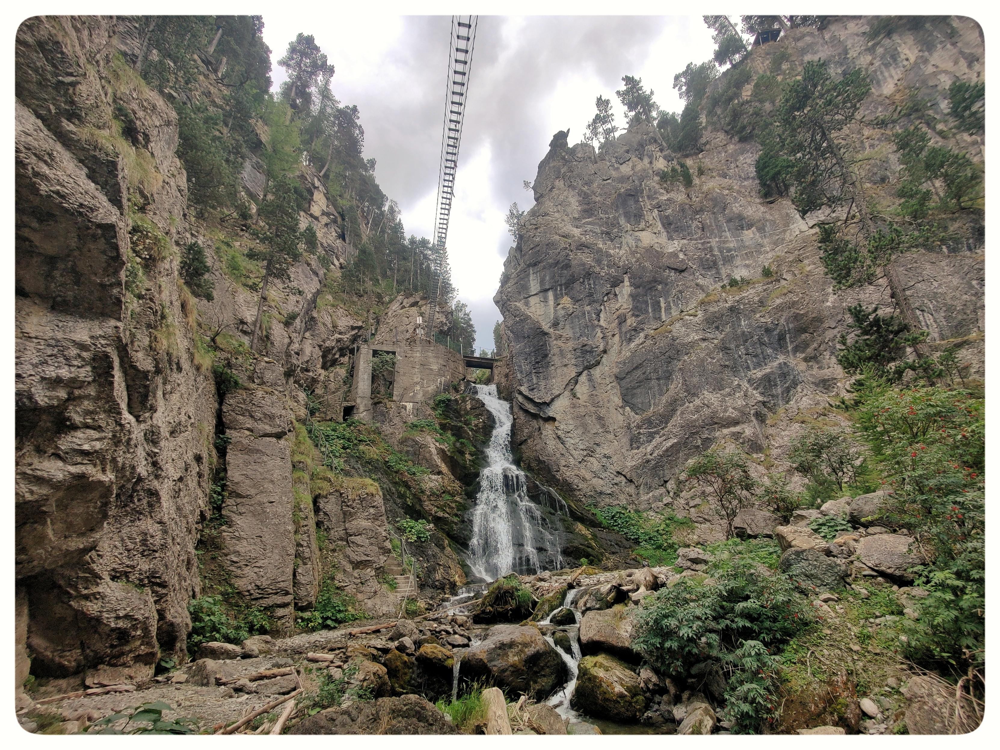 Scorcio di gole di montagna.
Il punto di vista è dal fondo della gola, dove scorre un torrente. Al centro, la cascata che scende dalle rocce. Sopra passano un ponte (normale) e un ponte tibetano. Le pareti di roccia scendono perpendicolari al terreno.