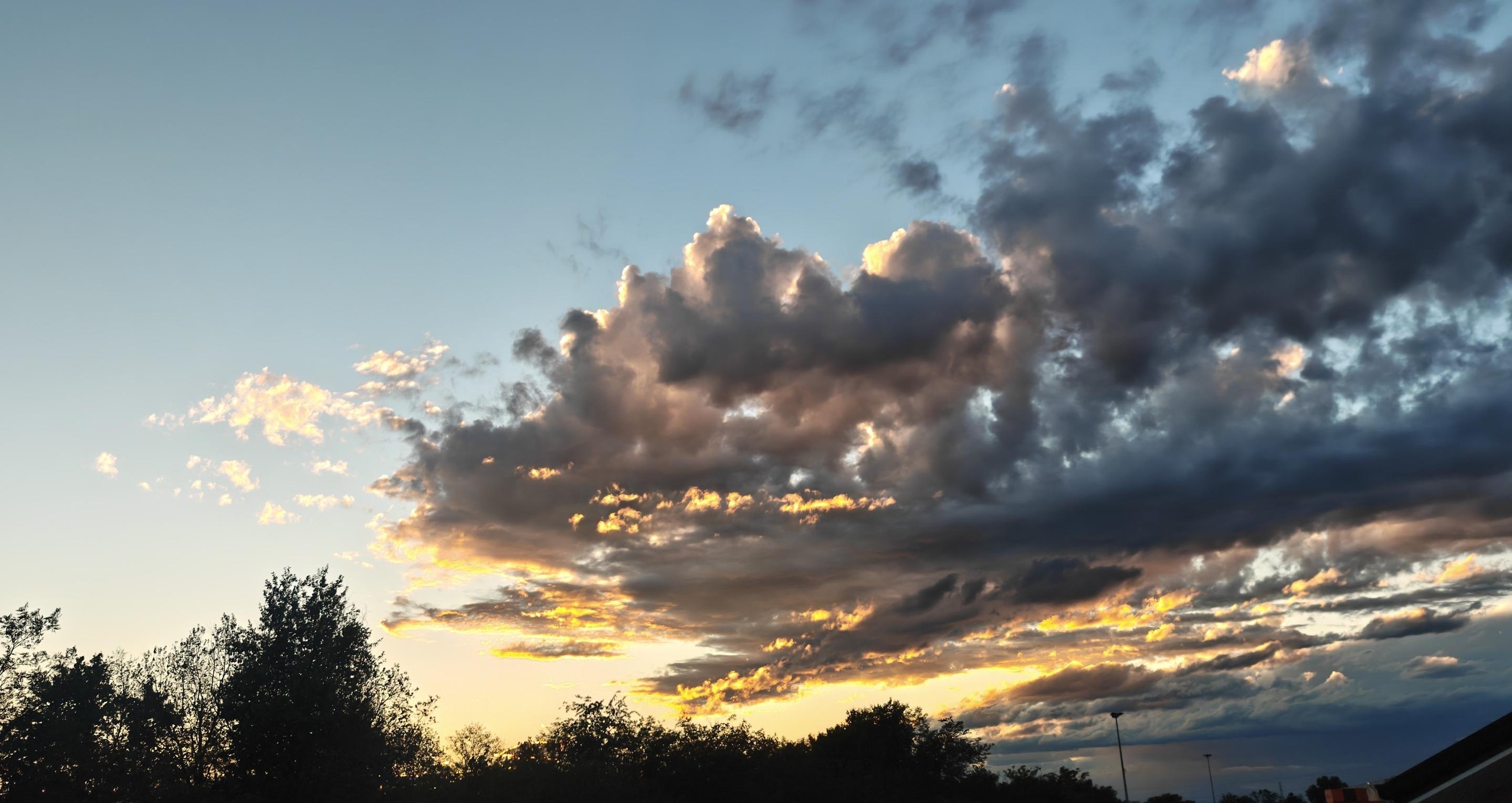 L'immagine mostra un tramonto suggestivo, caratterizzato da un forte contrasto tra luci e ombre.Il Cielo e le NuvoleColori caldi: Una luce dorata e arancione brilla all'orizzonte.Nuvole scure: Grandi nuvole grigie e bluastre coprono parte del cielo.Bordi illuminati: I profili delle nuvole riflettono i raggi solari con sfumature oro.Sfondo azzurro: La parte superiore del cielo sfuma in un azzurro chiaro e limpido.Il Paesaggio SottostanteSilhouette scure: La linea dell'orizzonte è segnata dai profili neri degli alberi.Elementi urbani: Sulla destra si intravedono i pali della luce e il tetto di un edificio.