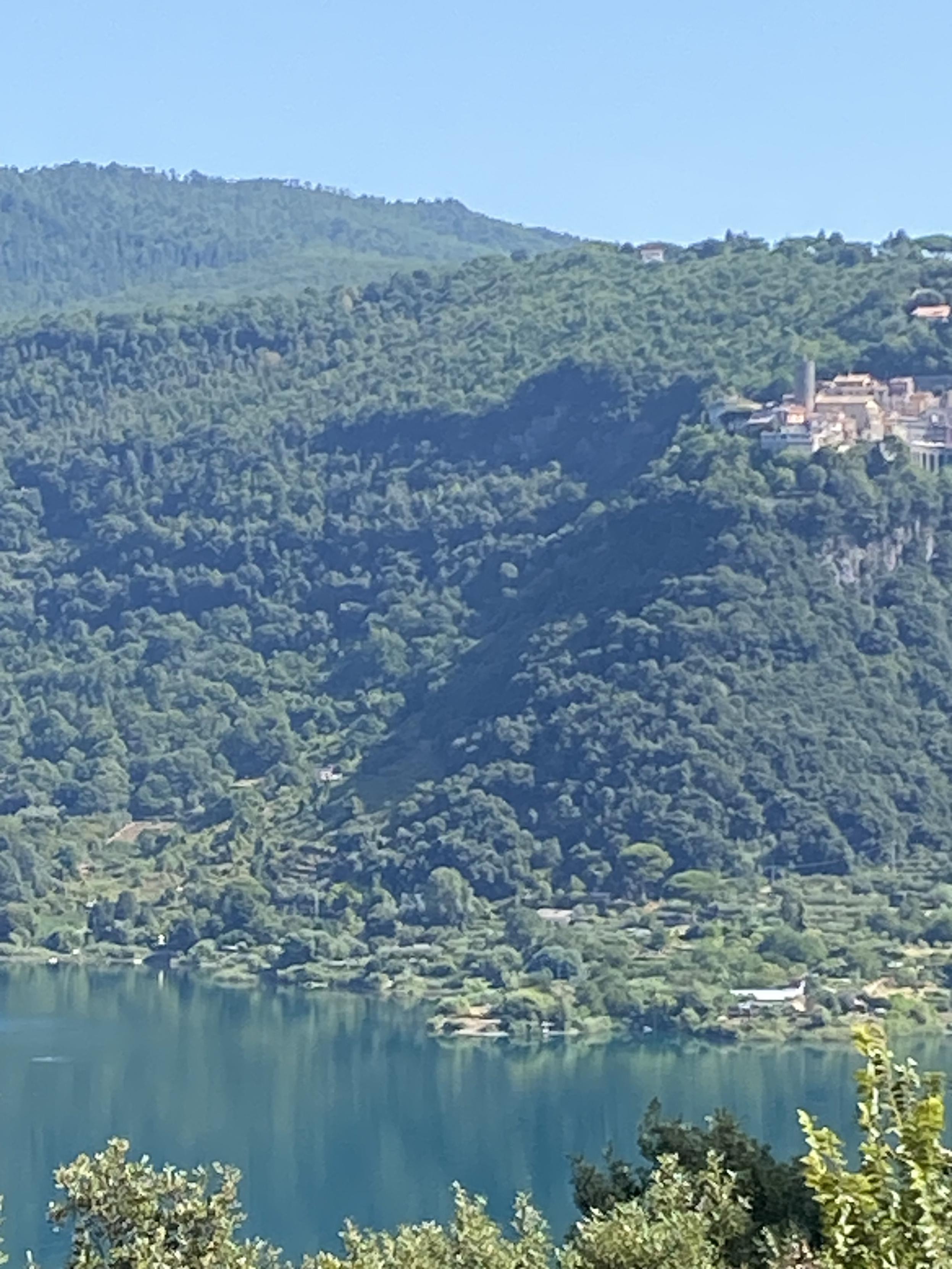 A tiny village sporting a round tower clings on a very steep wooded cliff that hands over a volcanic lake 
The sky is blue the waters very still and green/turquoise