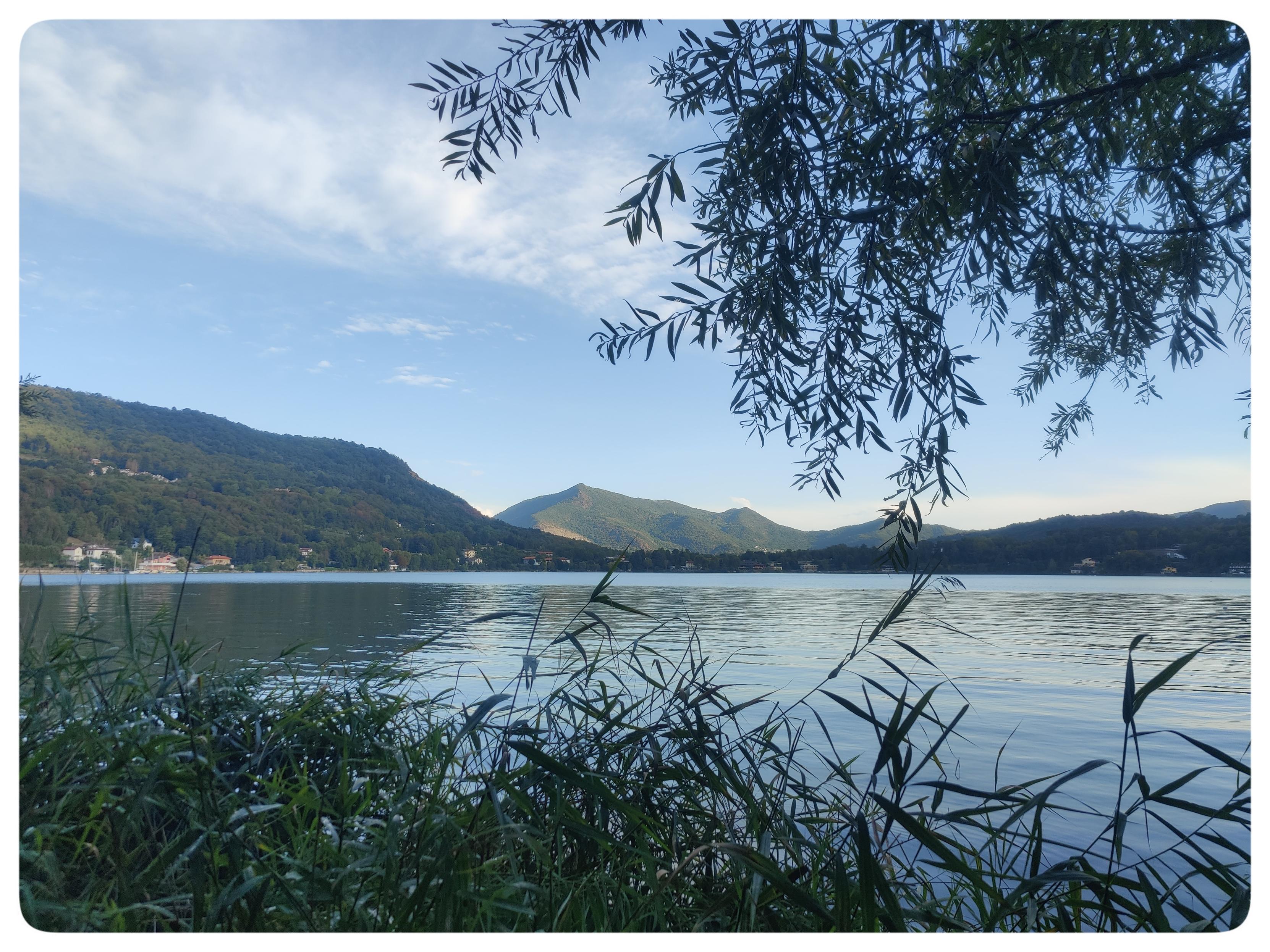 La versione intera della foto precedente.
Il lago al tramonto incorniciato dalle canne e dagli alberi che crescono sulle sue rive.
L'acqua riflette debolmente il cielo azzurro che inizia a tingersi di rosa. Una fila di montagne divide il lago dal cielo.