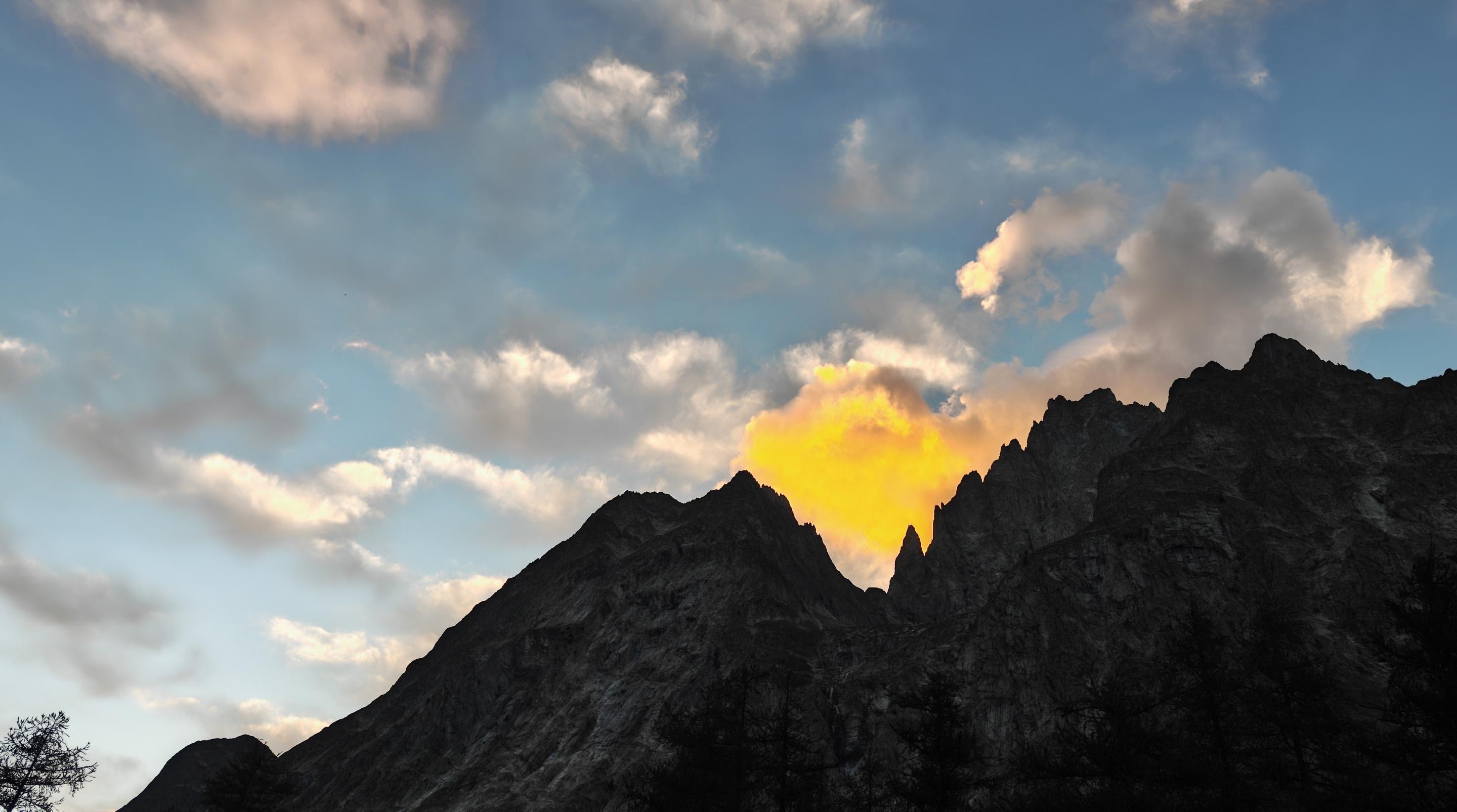 L'immagine cattura un suggestivo tramonto montano caratterizzato da un forte contrasto visivo.Ecco i dettagli principali della scena:Le montagne: Il profilo delle vette, frastagliato e roccioso, si staglia in controluce sul fondo. I rilievi sono scuri, quasi completamente in ombra.Il punto di luce: Dietro una sella tra le cime esplode una nube di colore giallo aranciato e dorato molto intenso. Sembra quasi che la montagna stia nascondendo un fuoco o che il sole stia calando proprio in quel punto esatto.Il cielo: La parte superiore è dominata da tonalità azzurre e sfumature di blu chiaro. È costellato da nuvole sparse, alcune più soffici e illuminate da tonalità rosate e dorate, altre più grigie e in ombra.La vegetazione: Nella parte inferiore si intravedono le sagome scure delle chiome di alcuni alberi (probabilmente larici o abeti), tipici del paesaggio alpino.