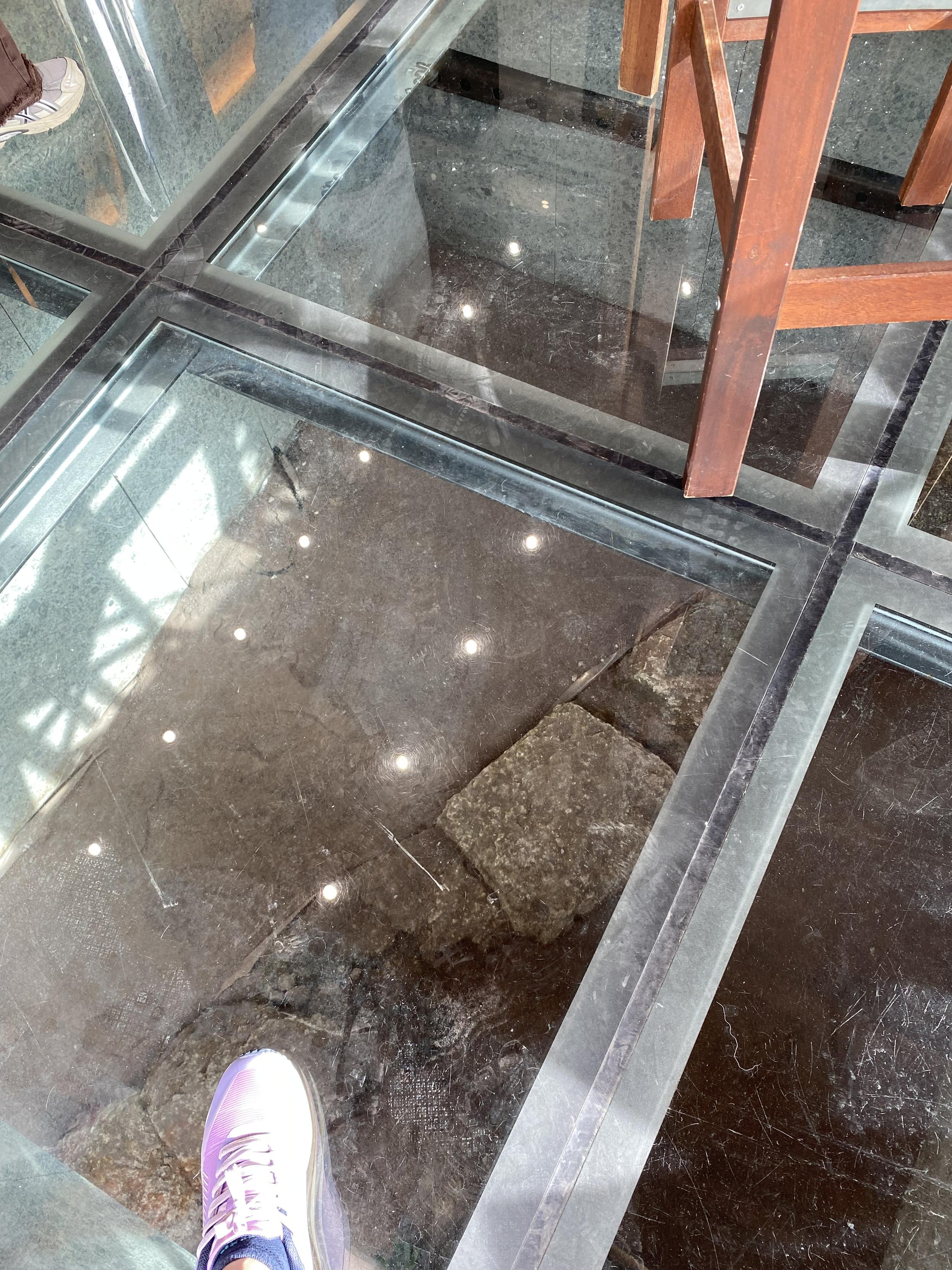 Old wall visible though a glass floor and my sneaker clad foot on it