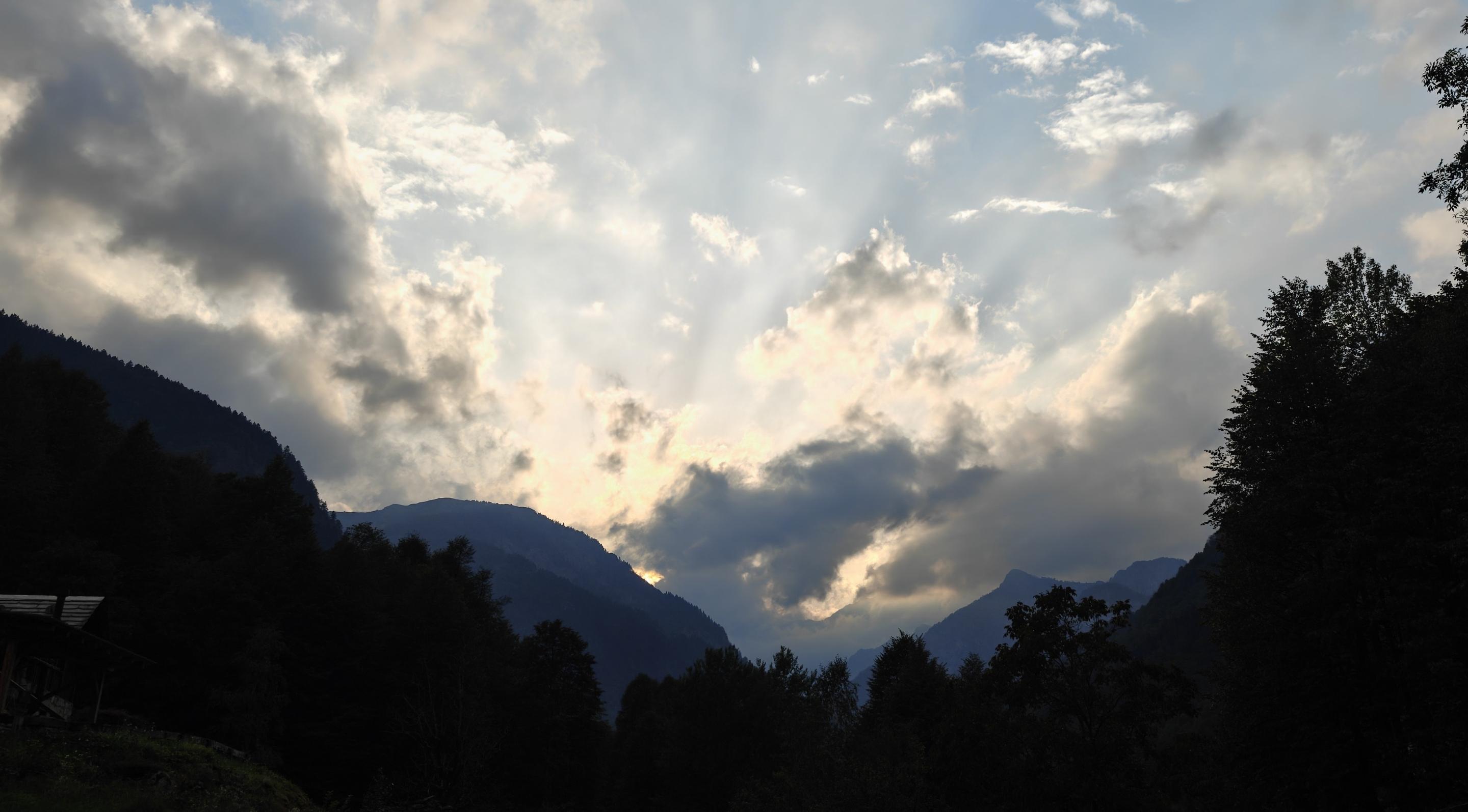 L'immagine mostra un affascinante tramonto tra le montagne, caratterizzato da un suggestivo gioco di luci e ombre creato dalle nuvole e dai rilievi alpini.Ecco i dettagli principali della scena:I raggi crepuscolari: Il sole, nascosto dietro lo strato nuvoloso centrale, proietta una spettacolare raggiera di luce (chiamata anche "dita di Dio") che si estende verso l'alto, squarciando il cielo azzurro e illuminando le nuvole circostanti con sfumature dorate e bianche.Le montagne e la vegetazione: I profili dei rilievi montuosi e la fitta foresta di alberi in primo piano appaiono completamente in ombra, creando una netta silhouette scura che incornicia il paesaggio e accentua il contrasto con la luminosità del cielo.Il cielo e le nuvole: Il cielo sovrastante è in gran parte sereno e di una tonalità azzurro chiaro, parzialmente coperto da nuvole sparse. Le nuvole più basse, vicine al sole calante, mostrano bordi luminosi e caldi, mentre quelle più alte rimangono di una tonalità grigio-azzur