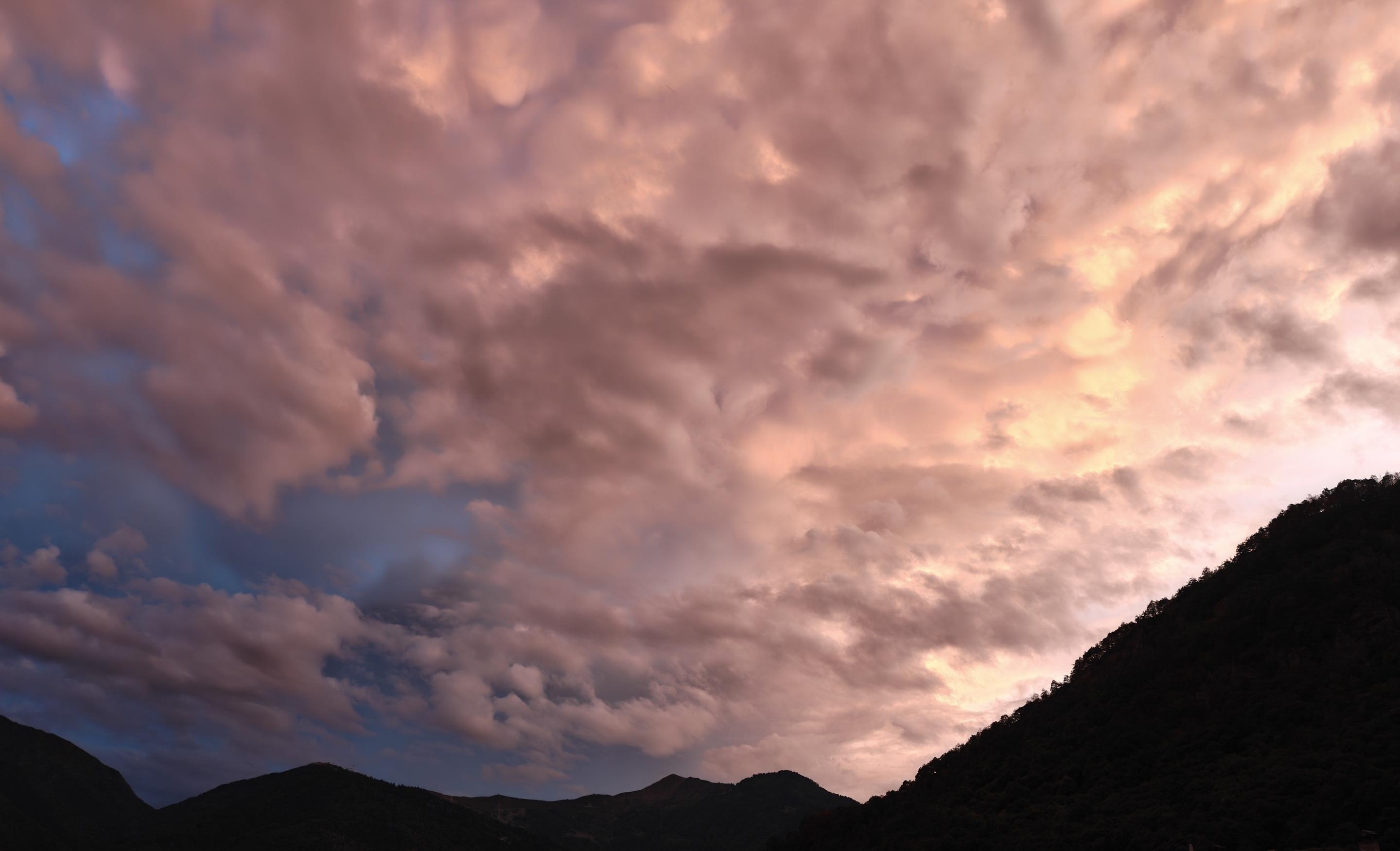 Un tramonto suggestivo e dinamico colora il cielo sopra un profilo montuoso scuro. La scena è dominata da una fitta coltre di nuvole frastagliate e stratificate, che catturano gli ultimi raggi di sole creando un ricco contrasto cromatico.Le caratteristiche principali dell'immagine includono:I colori del cielo: Le nuvole sono tinte di sfumature calde che vanno dal rosa intenso al pesca e all'arancione dorato sulla destra, dove la luce solare è più forte. Sulla sinistra, il cielo vira verso tonalità più fredde, rivelando squarci di azzurro e blu profondo.La consistenza delle nuvole: La copertura nuvolosa è densa e mossa, con cumuli che creano un gioco di luci e ombre, dando volume e un senso di movimento a tutta la composizione.Il paesaggio sottostante: In basso si staglia la silhouette scura e netta delle montagne, che fa da contrasto alla luminosità e alla vivacità del cielo sovrastante.