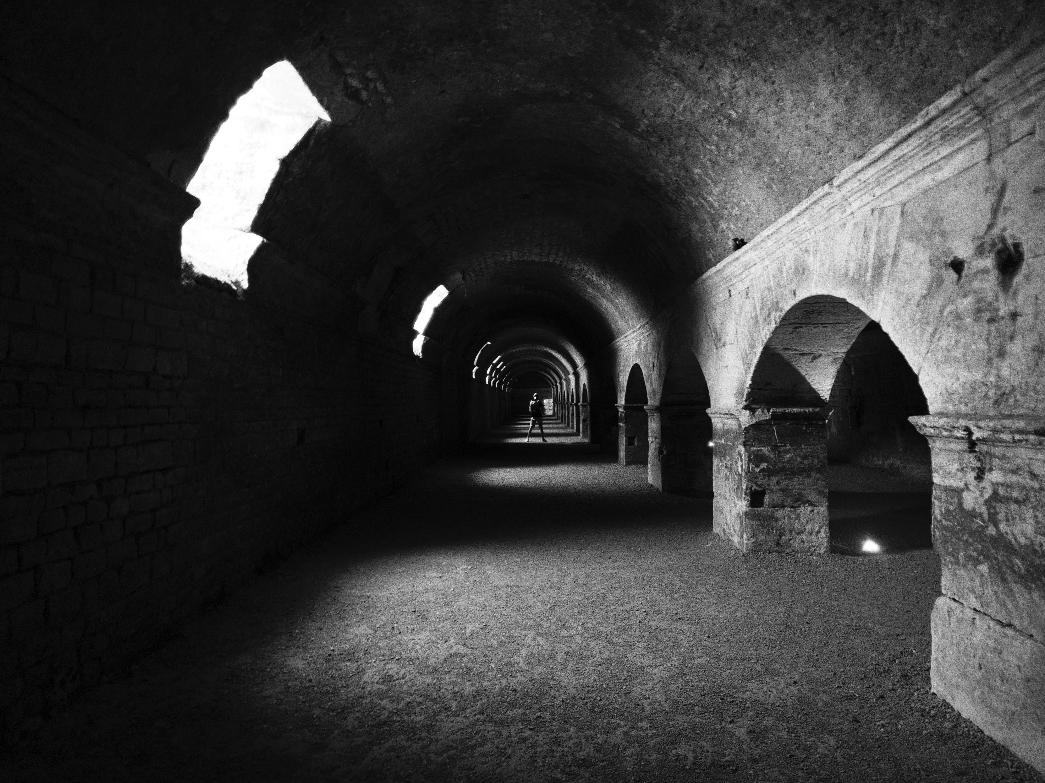 La foto mostra un lungo corridoio sotterraneo in pietra, illuminato da piccole aperture nella volta a botte, che lasciano entrare fasci di luce naturale. L’ambiente ha un aspetto antico, con archi e pilastri che sostengono il soffitto. Il pavimento è di terra battuta e l’intero corridoio trasmette un’atmosfera misteriosa e silenziosa. In lontananza si intravede una figura umana, piccola rispetto alla vastità del passaggio, che accentua la profondità e la prospettiva della scena.