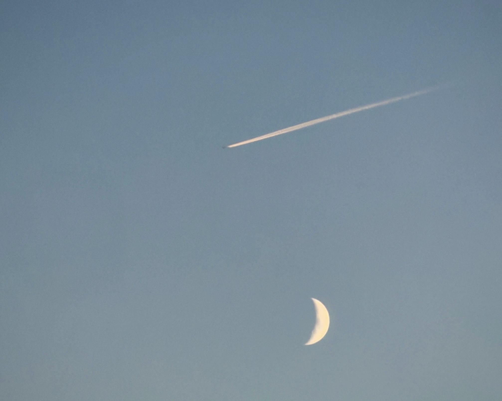 Luna crescente e un aereo con scia nel cielo sereno.
