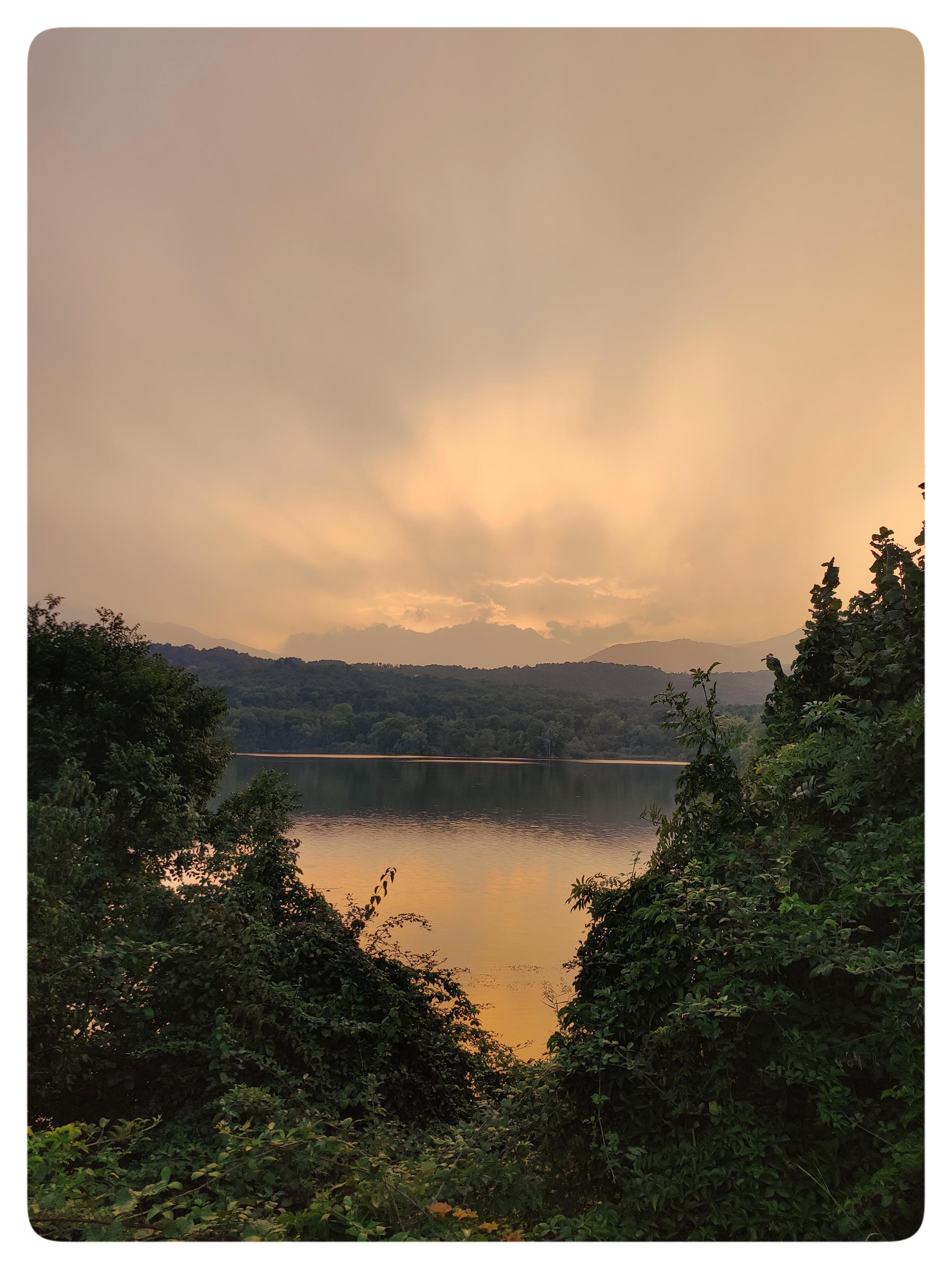 Tramonto sul lago.
Le acque del lago riflettono la luce del tramonto. La scena è incorniciata dalla vegetazione che cresce sulle sponde. Il sole è nascosto dalle nuvole e solo la lice filtra e illumina la parte alta della foto, creando l'effetto di raggi solari.