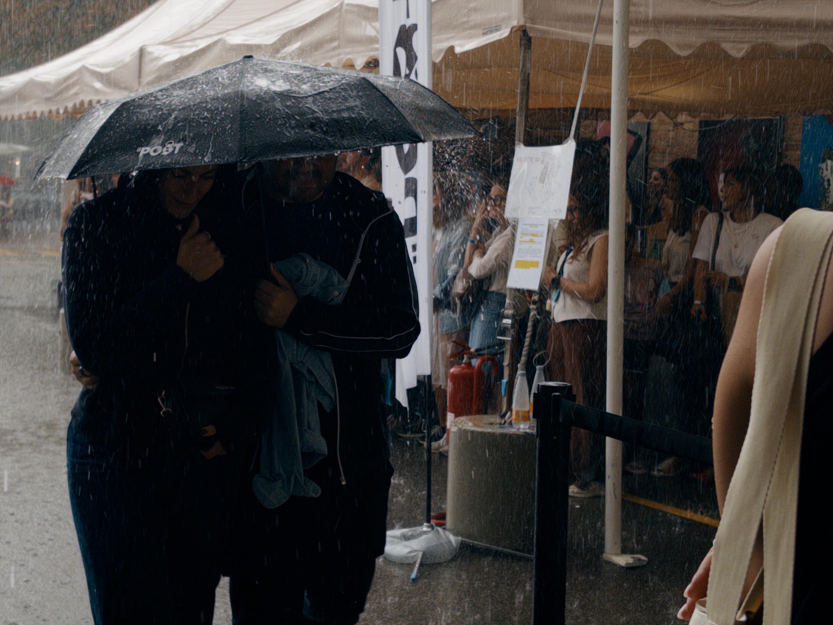 La foto mostra due persone che camminano strette sotto un grande ombrello nero mentre piove intensamente. Sullo sfondo ci sono tende da mercato o stand con persone riparate sotto i tendoni, alcune delle quali osservano verso l’esterno. L’acqua scende copiosa, creando schizzi evidenti sul terreno bagnato. L’atmosfera suggerisce un acquazzone improvviso durante un evento all’aperto.