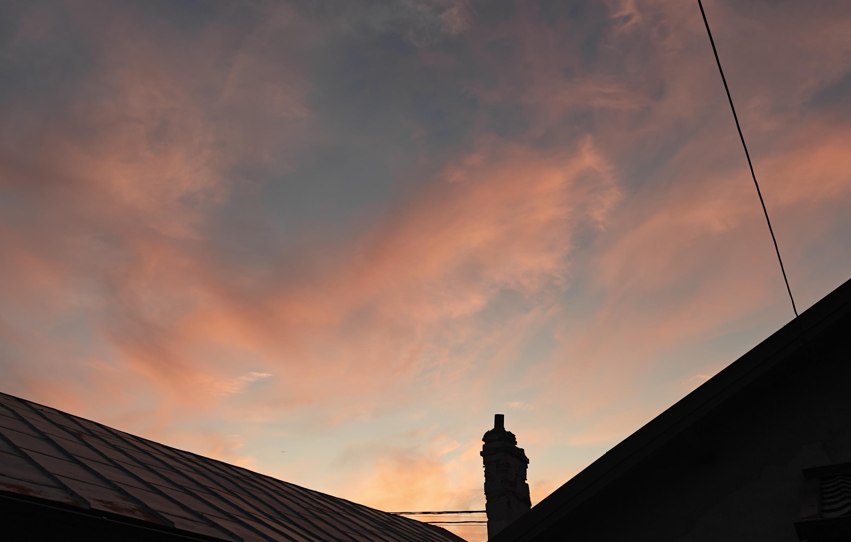 L'immagine mostra un suggestivo tramonto caratterizzato da sfumature calde e delicate, catturato da una prospettiva urbana o domestica.Ecco i dettagli principali della scena:Il cielo e le nuvole: Il cielo è attraversato da nuvole leggere e sfilacciate, simili a cirri, che si tingono di tonalità rosa, salmone e arancione dorato. Sullo sfondo, nelle zone meno illuminate dal sole calante, il cielo vira verso un grigio-azzurro tenue e un blu polvere, creando un contrasto morbido.Le silhouette in primo piano: Nella parte inferiore dell'inquadratura si stagliano le sagome scure e in controluce di alcuni tetti. A sinistra si nota un tetto inclinato in lamiera o metallo, mentre a destra la linea scura di un altro edificio incornicia la scena.Elementi verticali: Al centro, tra i due tetti, emerge la silhouette slanciata di un comignolo antico o decorato. Nell'angolo in alto a destra, invece, un cavo elettrico taglia diagonalmente il cielo in modo netto.