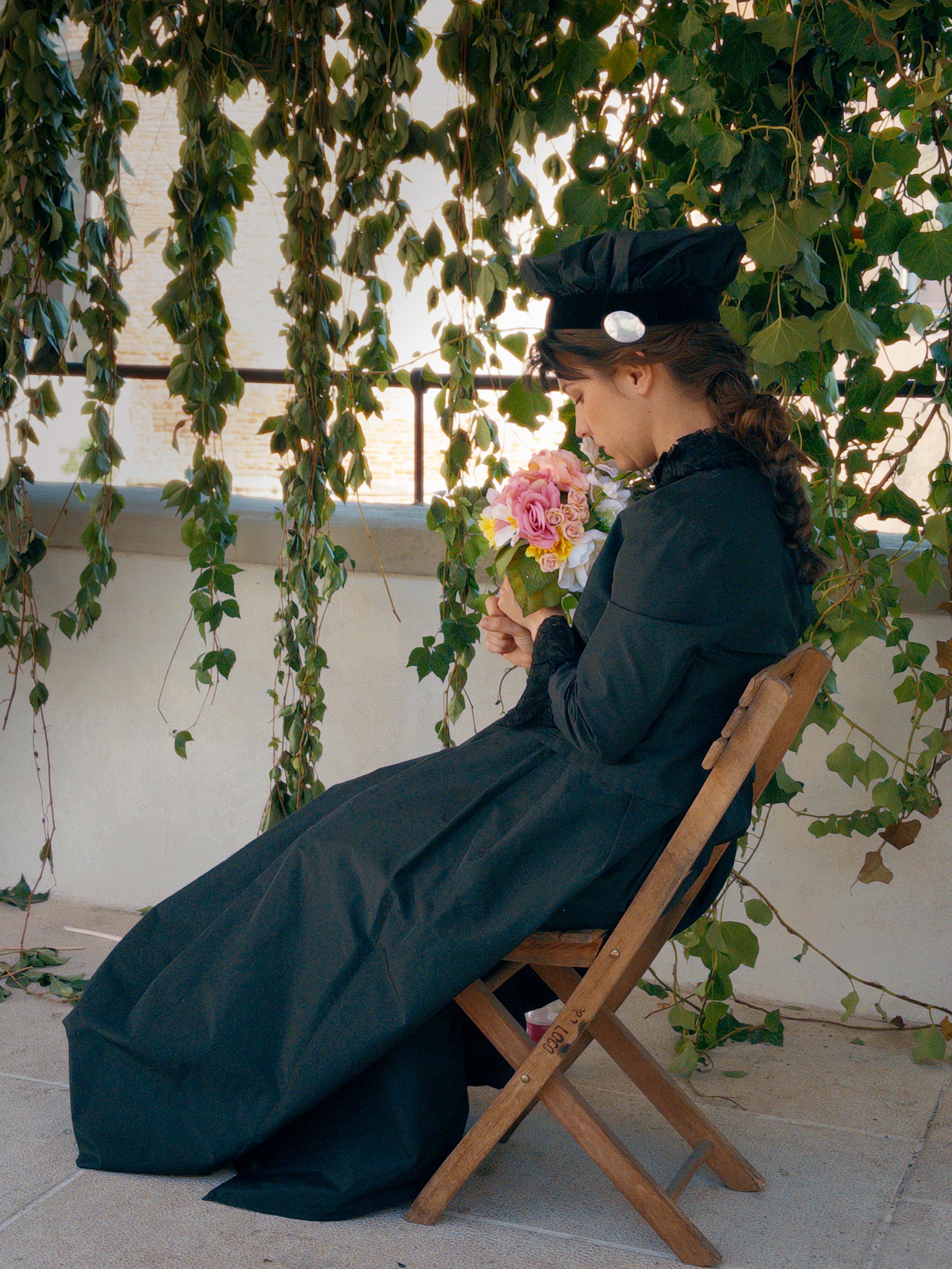 La foto mostra una donna seduta su una sedia pieghevole di legno, di profilo, con lo sguardo rivolto verso un piccolo bouquet di fiori rosa e gialli che tiene tra le mani. Indossa un lungo abito nero in stile retrò e un cappello scuro con un ornamento chiaro. Sullo sfondo ci sono rampicanti verdi che scendono da un pergolato, filtrando la luce naturale e creando un’atmosfera tranquilla e malinconica.