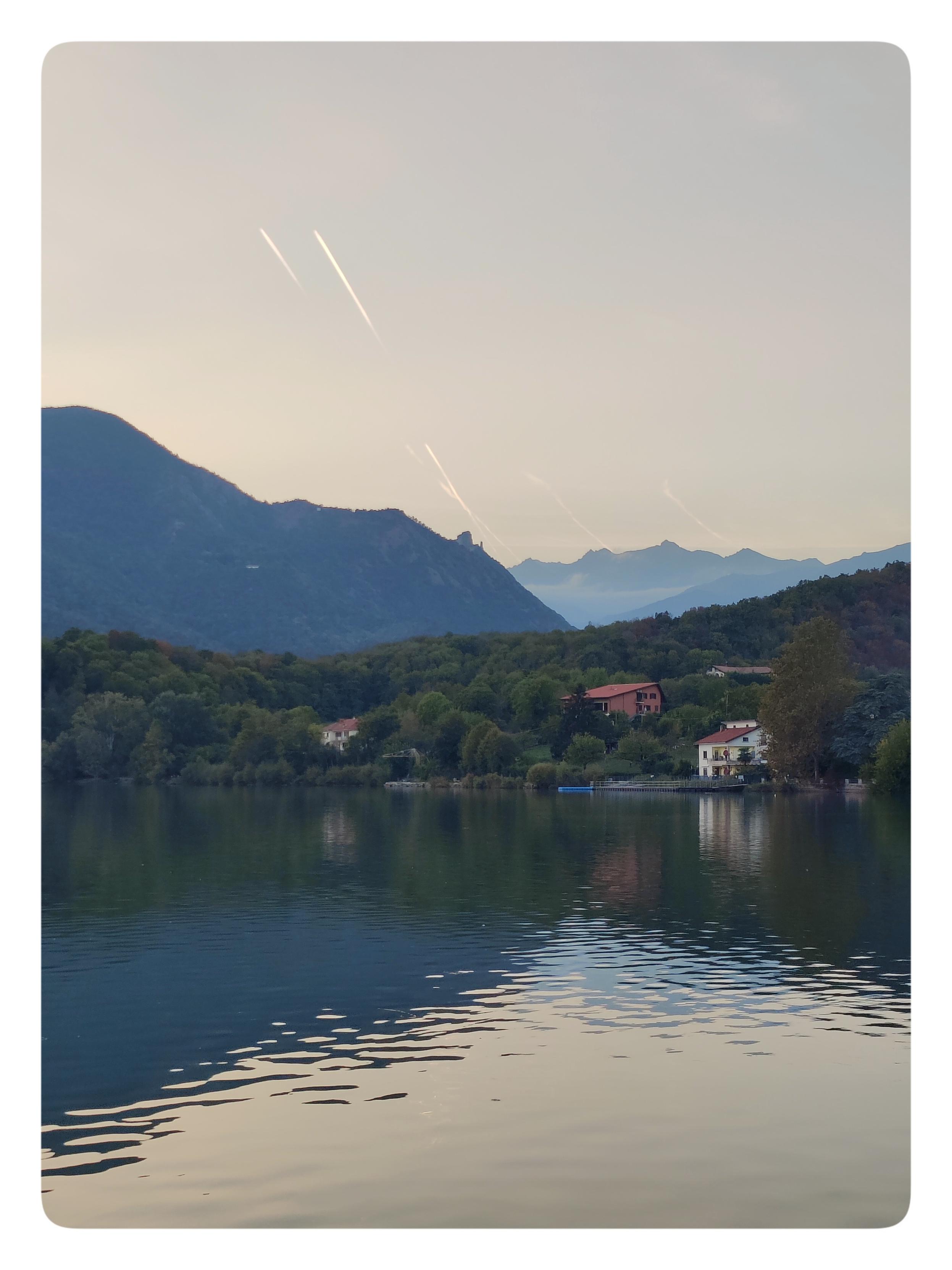 La Sacra.
Il lago è un po' mosso. L'acqua riflette un indizio di montagne.
Le montagne più vicine nascondono alcune case tra i loro alberi.
Le montagne più lontane sono scure. Su una di esse si staglia il profilo della Sacra di San Michele.
Il cielo è solcato dalle scie di alcuni aerei.