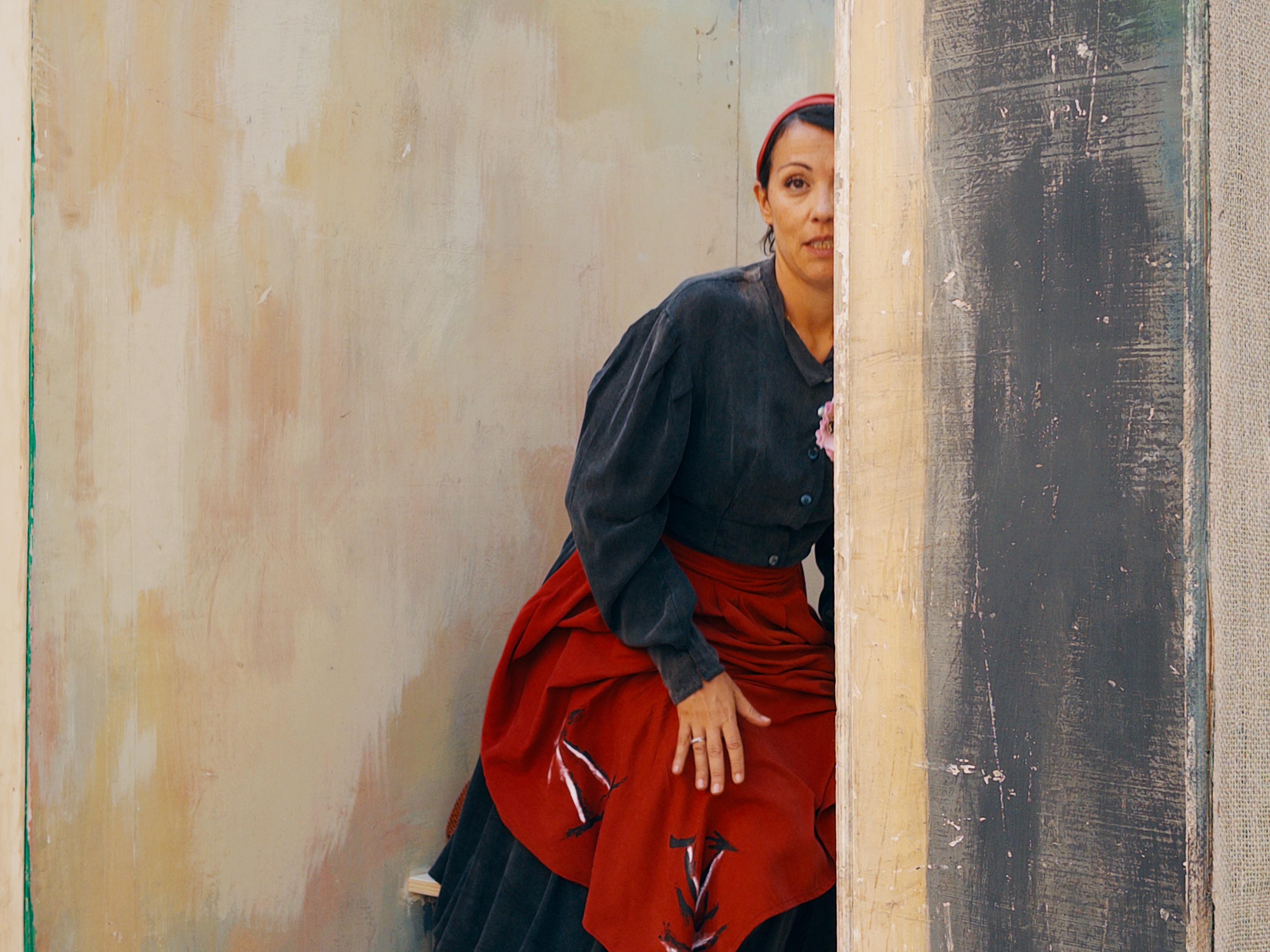 La foto mostra una donna che sbircia da dietro un angolo, parzialmente nascosta. Indossa una camicetta scura e una lunga gonna nera con un grembiule rosso sopra, decorato con piccole linee bianche. Ha una fascia rossa tra i capelli. Sullo sfondo si vedono pareti dall’aspetto rustico e pittorico, con colori chiari e sfumati, che creano un’atmosfera semplice e tradizionale.