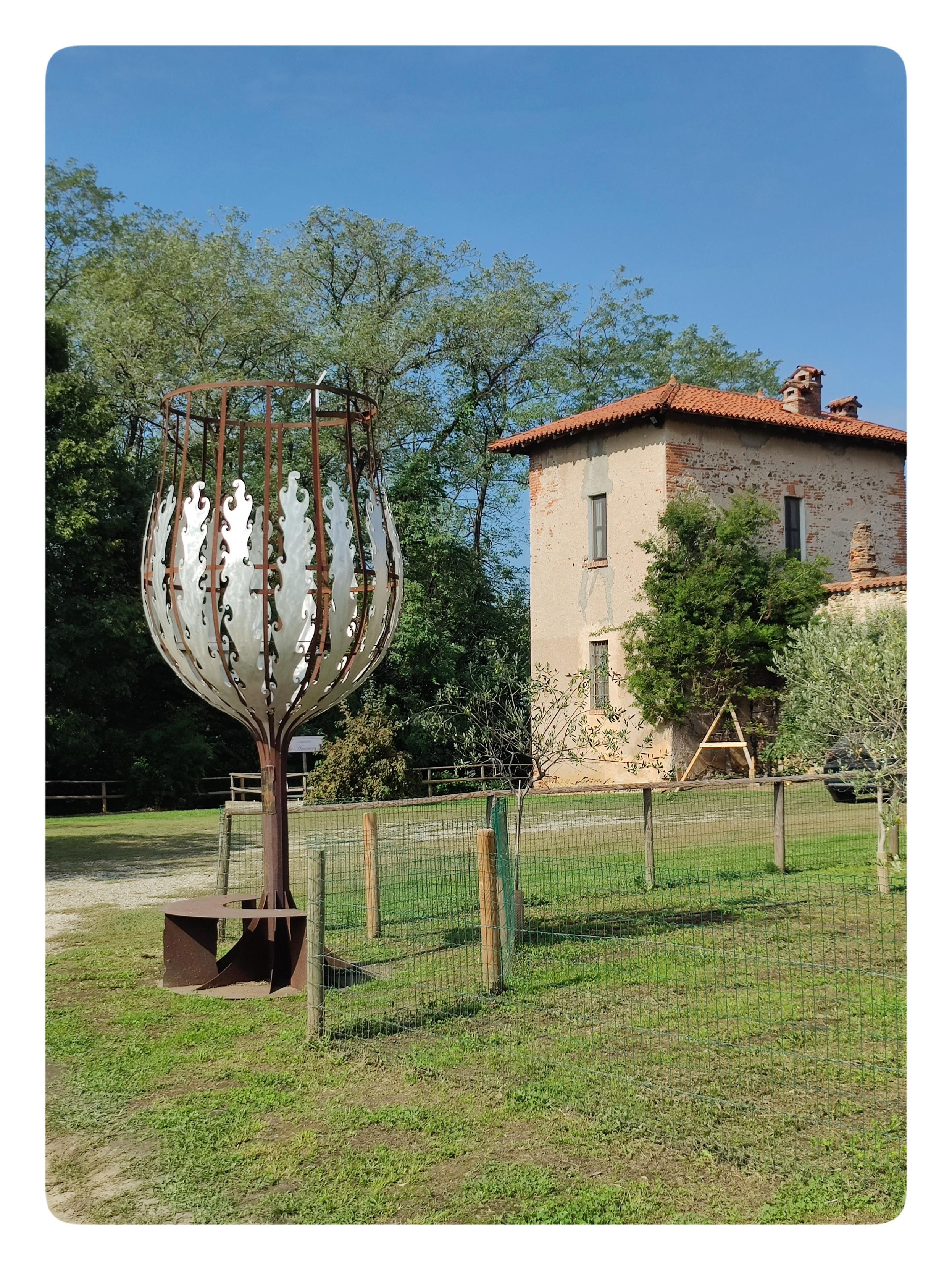 Il Calice Gigante numero 12. Davanti alla Cascina Cavenago (che, indovinate un po', è stata un castello!). Il calice ha una struttura in metallo. La coppa del calice è decorata in metallo chiaro, che riflette la luce del sole. A lato si vede uno dei torrioni del castello, in parte intonacato, in parte con i mattoni a vista. Oggi funge da ingresso alla tenuta.