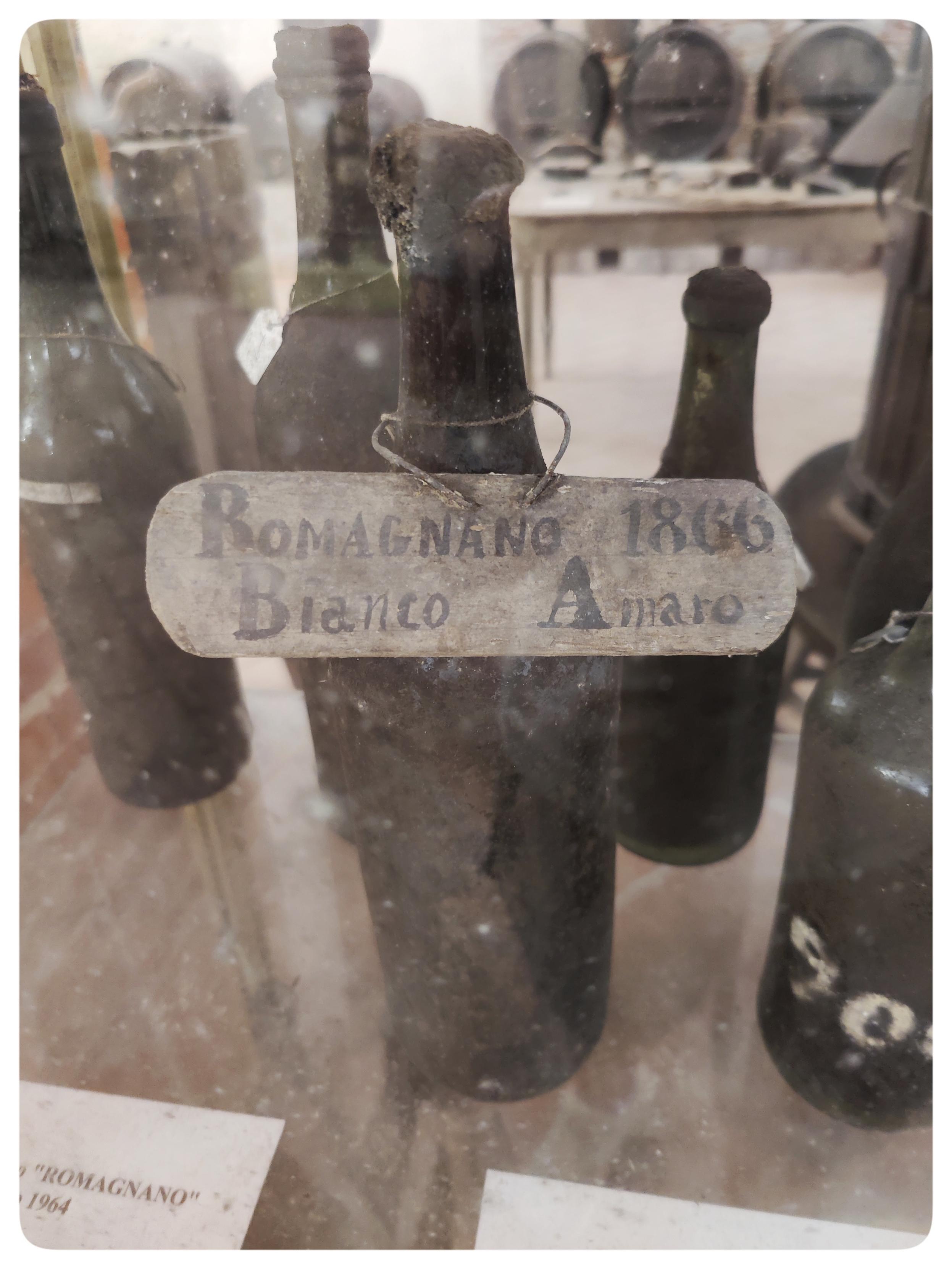 Bottiglia di Romagnano Bianco Amaro datata 1866. Si trova in una delle teche del MEV. Il corpo della bottiglia è scuro, il tappo annerito da tempo e umidità. Intorno, si vedono altre bottiglie antiche all'interno della vetrina.