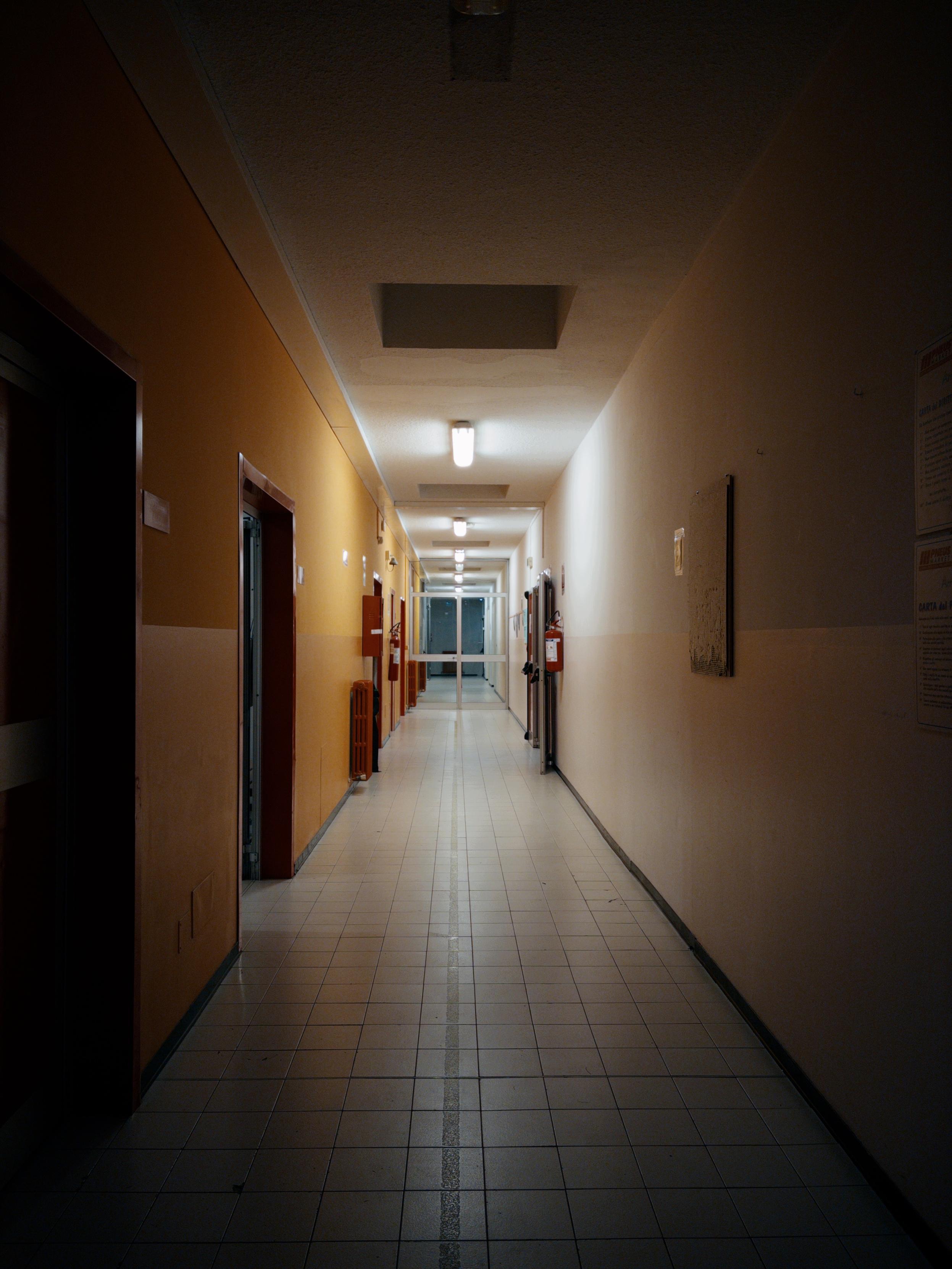 L’immagine mostra un lungo corridoio interno, illuminato da luci al soffitto che creano un’illuminazione uniforme. Le pareti sono di due colori diversi: la parte sinistra in giallo ocra e la destra in bianco. Il pavimento è piastrellato di colore chiaro con una sottile linea centrale scura che corre lungo il corridoio. Sul lato sinistro si notano porte scure con targhette, mentre a destra ci sono un quadro e alcuni avvisi affissi al muro. In fondo al corridoio si intravede una porta a vetri che sembra condurre verso l’esterno o un’altra sezione dell’edificio.