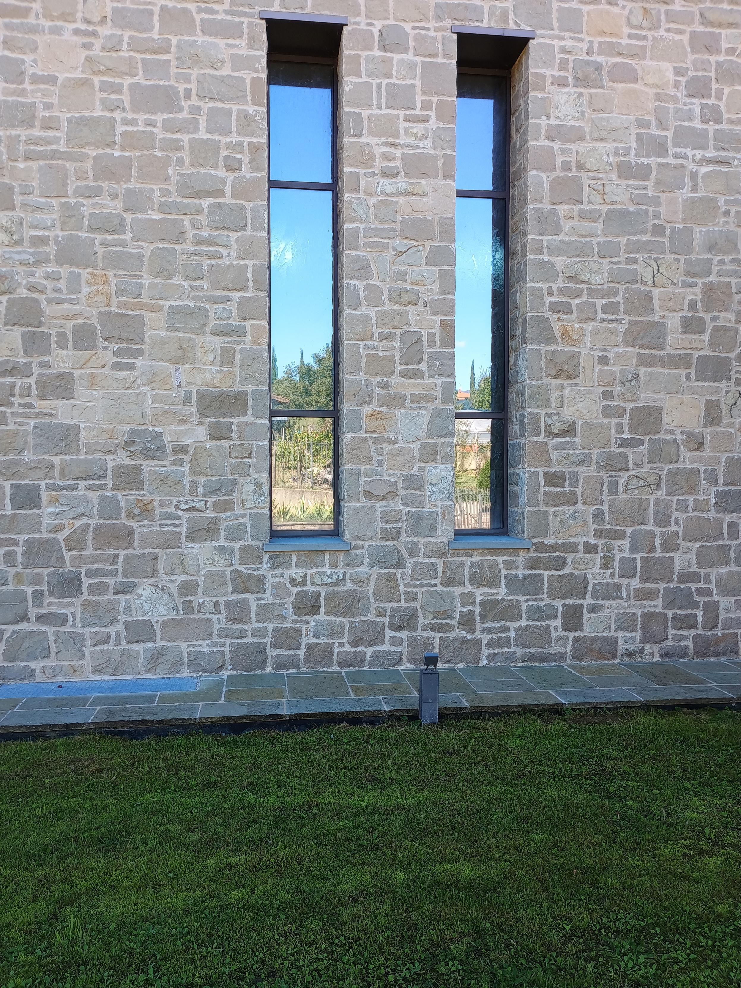 Una fotografia di una parete in pietra con due finestre alte e strette. La parete è composta da pietre irregolari di varie tonalità di grigio e beige. beige. Le finestre hanno cornici scure e riflettono un cielo azzurro e della vegetazione lontana. Alla base della parete si trova un bordo di pietra piatta con una piccola lampada rettangolare scura. In primo piano è visibile un prato di erba verde.