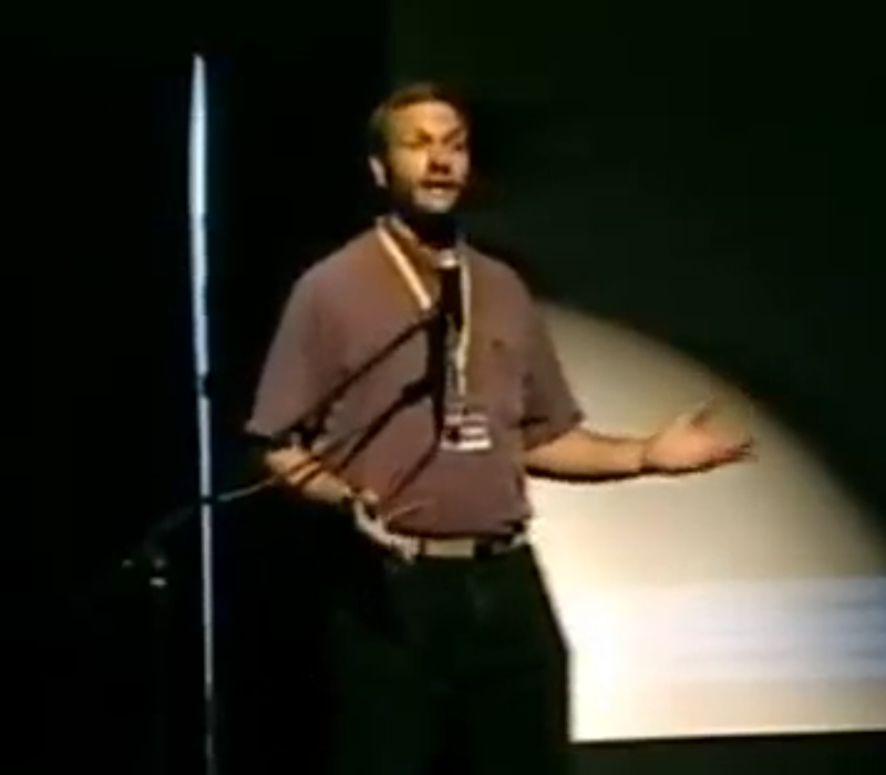 Photo of Greg Newby speaking on stage in a blurry screenshot from old low-fi video of his H2K panel "The Hackers Code." He wears a plain t-shirt and trousers and is gesturing expansively as he speaks.