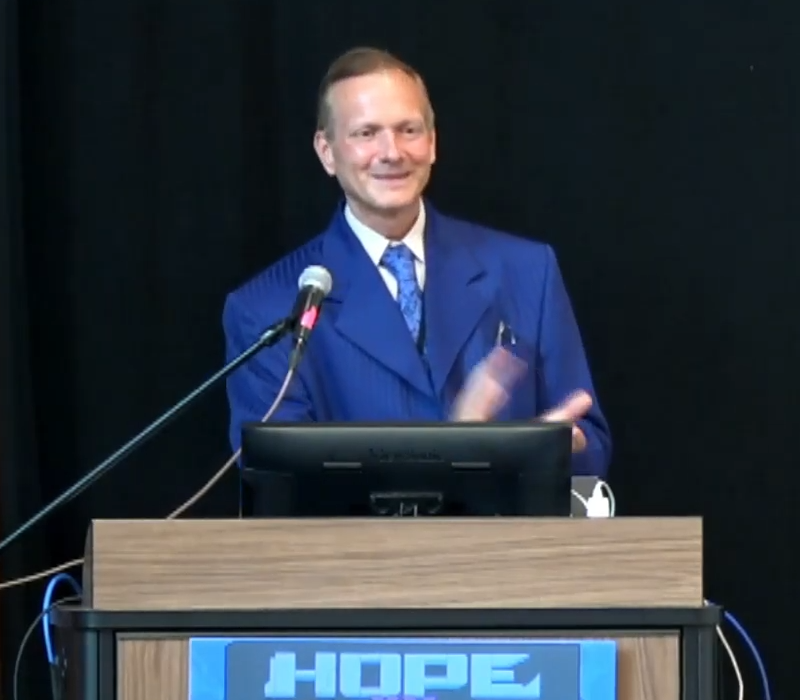 Greg Newby at the lectern at HOPE closing ceremonies. He is dressed in his trademark blue pinstriped zoot suit which became his trademark through most of HOPE history, and he smiles as he applauds the crowd.