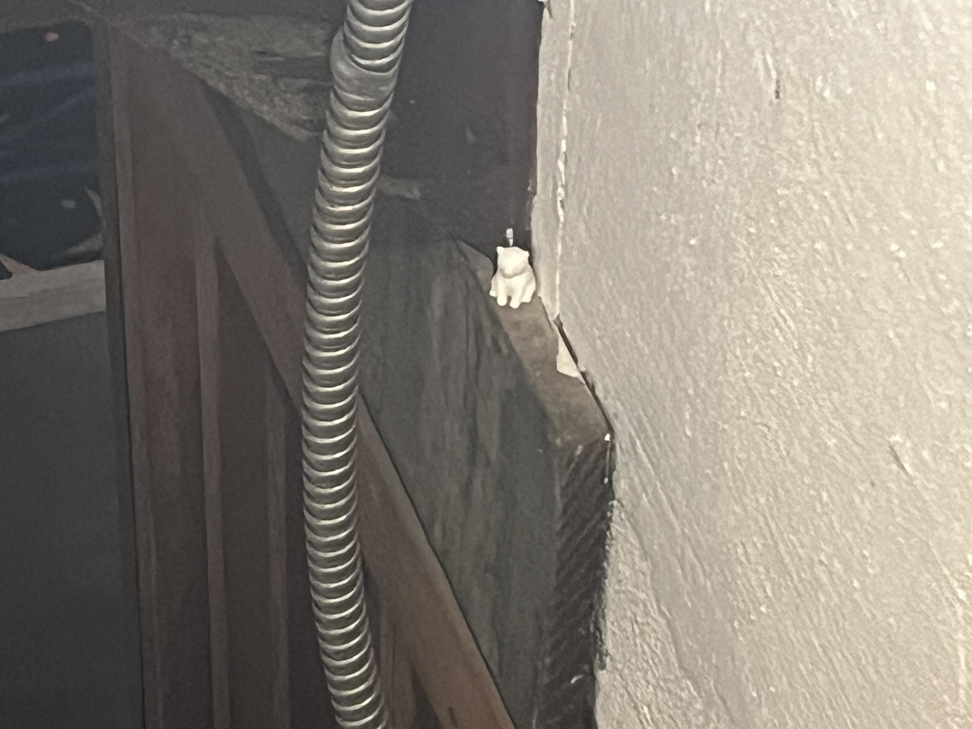 A small white figurine of a cat sits on a narrow wooden ledge near a wall. A metal conduit runs alongside it. The background is dimly lit.