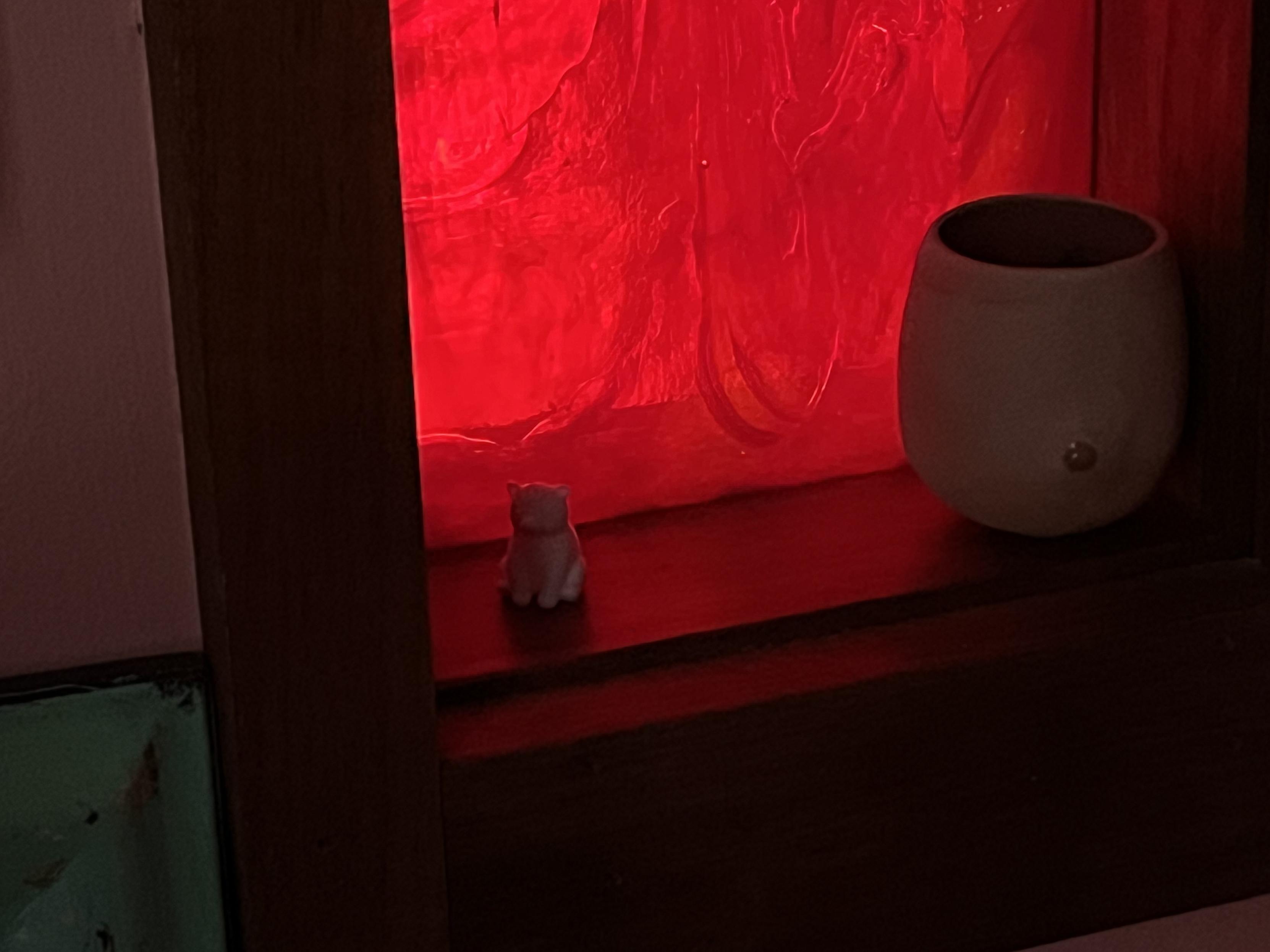 A small white cat figurine sits on a shelf next to a round, beige pottery vase. The background is illuminated with a red glow, enhancing the warm ambiance