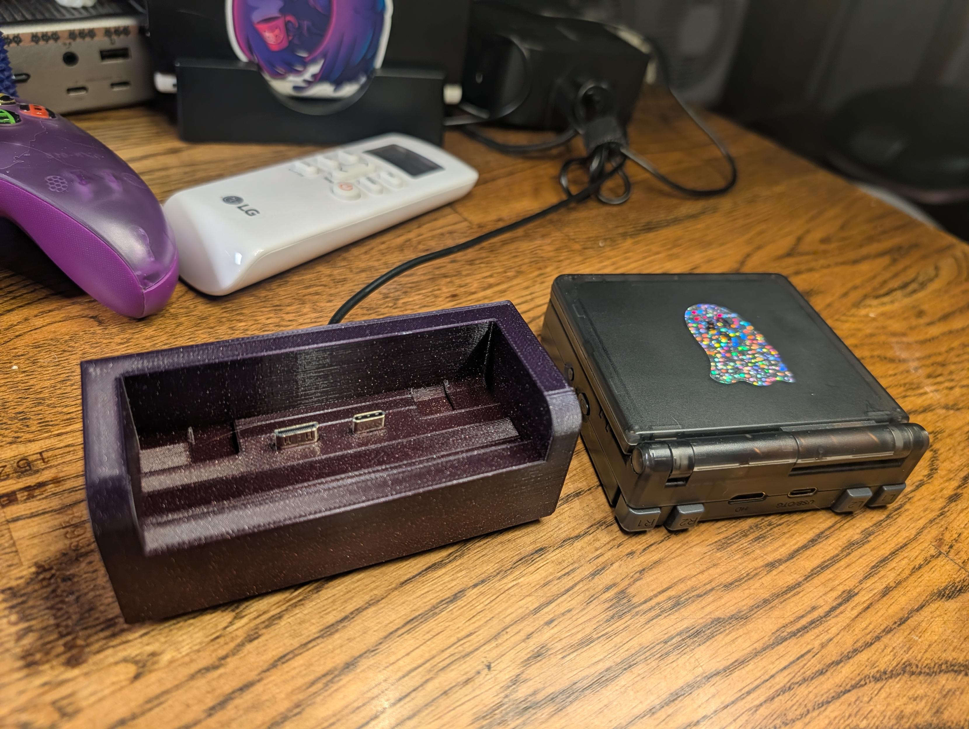 A black handheld gaming console with a ghost sticker sits next to its custom, 3D-printed purple charging dock on a wooden desk.