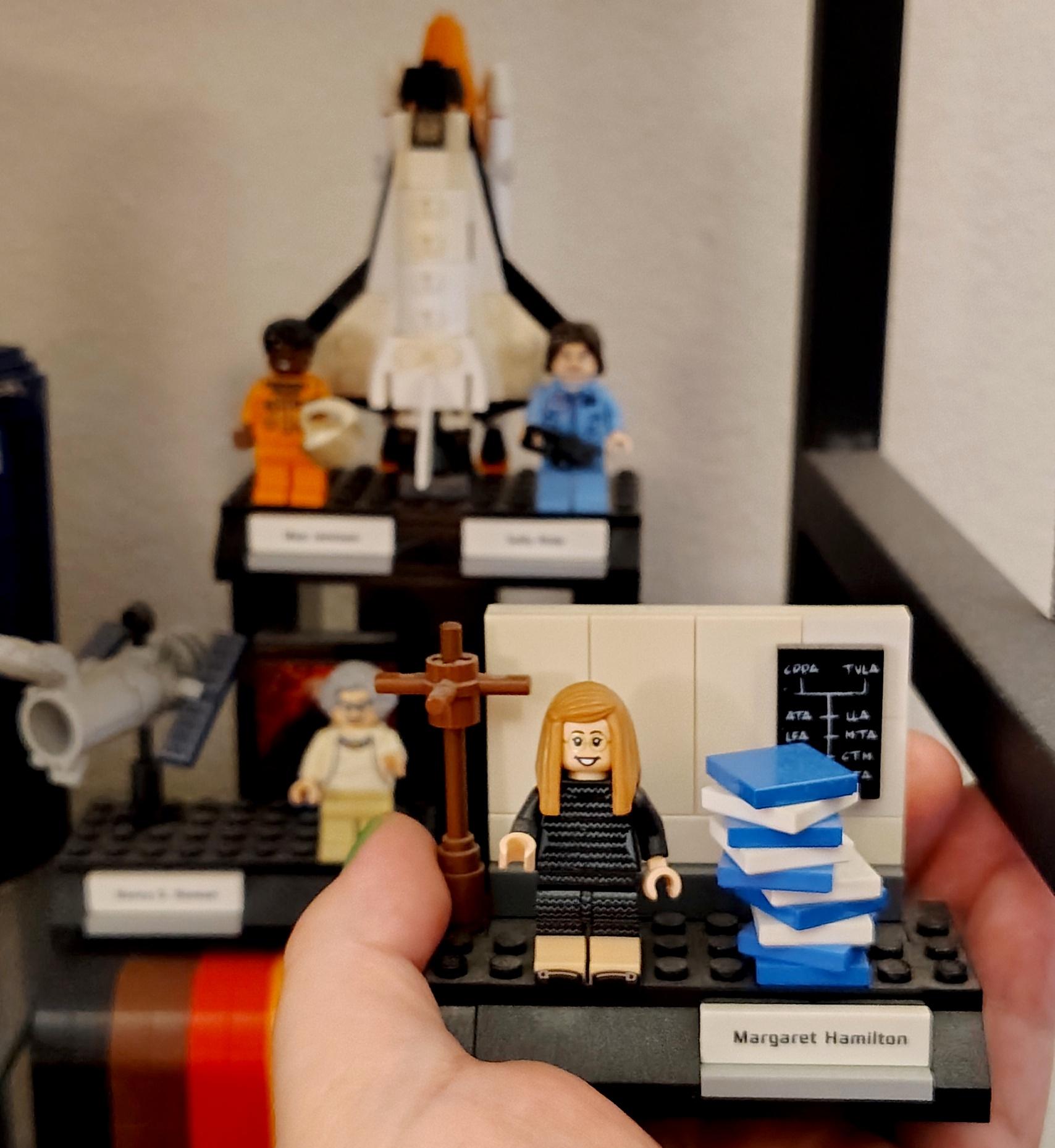A Lego display of Hamilton based on the previous photo, held aloft by the Undersigned in front of the rest of the "Women of NASA" Lego set it comes from which contains displays of Mae Jamison, Sally Ride, and Nancy Grace Roman.