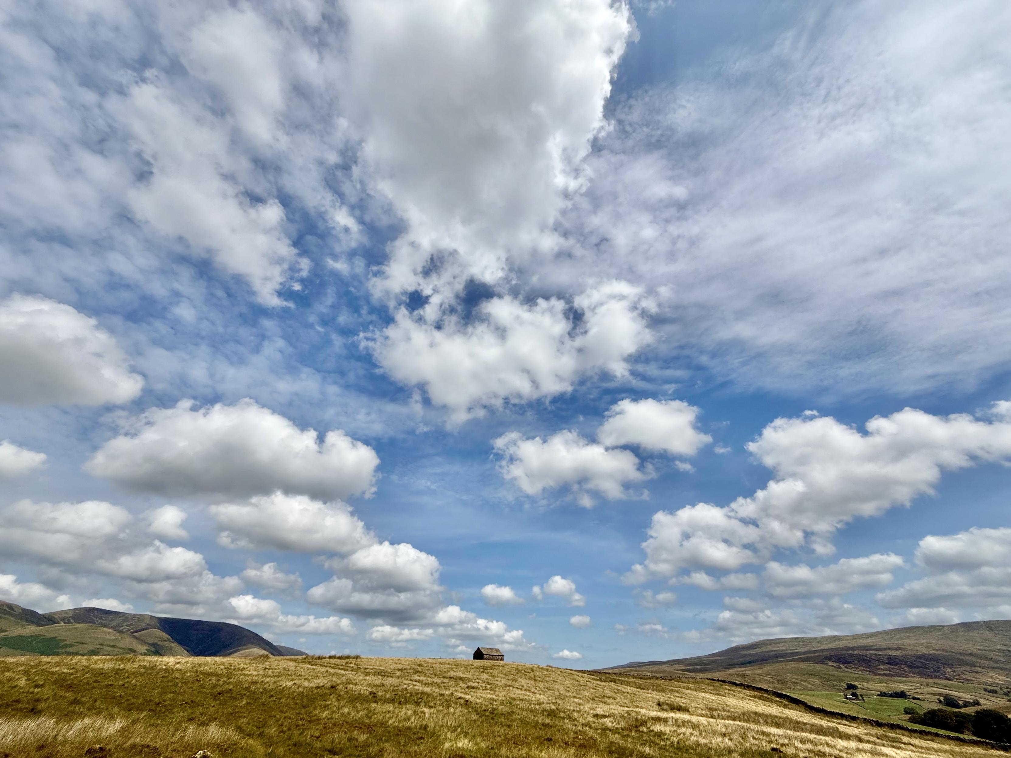 Stone barn in the middle of moorland under a sky full of fluffy clouds.