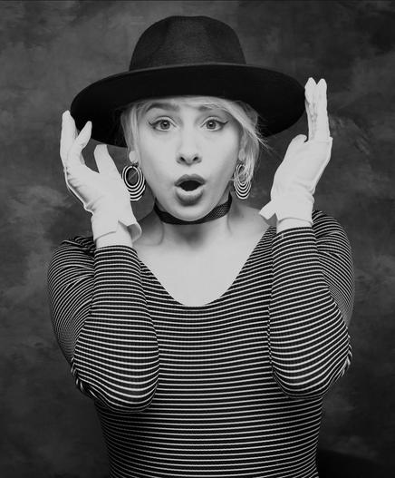 Olga Solo, a model, actor, performer, is dressed as a mime with many stripes, gloved hands up, and an exaggerated look of surprise on her face. In mono.
The black hat and striped earings add nothing but good.