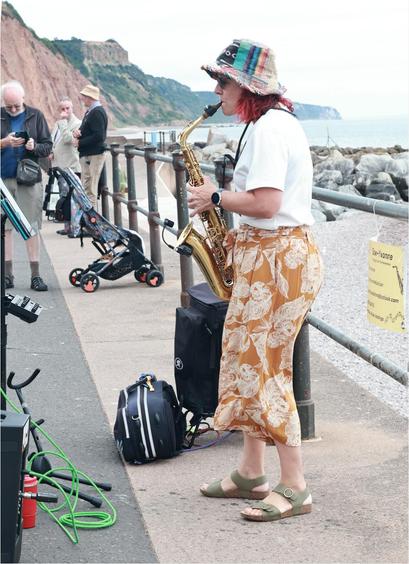 A saxophonist called Yvonne stands facing left, distal are the sandstone cliffs of the Jurassic Coast beyond Sidmouth and some sea and pebbles. Behind her isthe beach and a notice giving some details of her. To camera left, in front of her, some busker equipment including a Sax stand. There are 3 bystanders visible, not visibly paying attention.