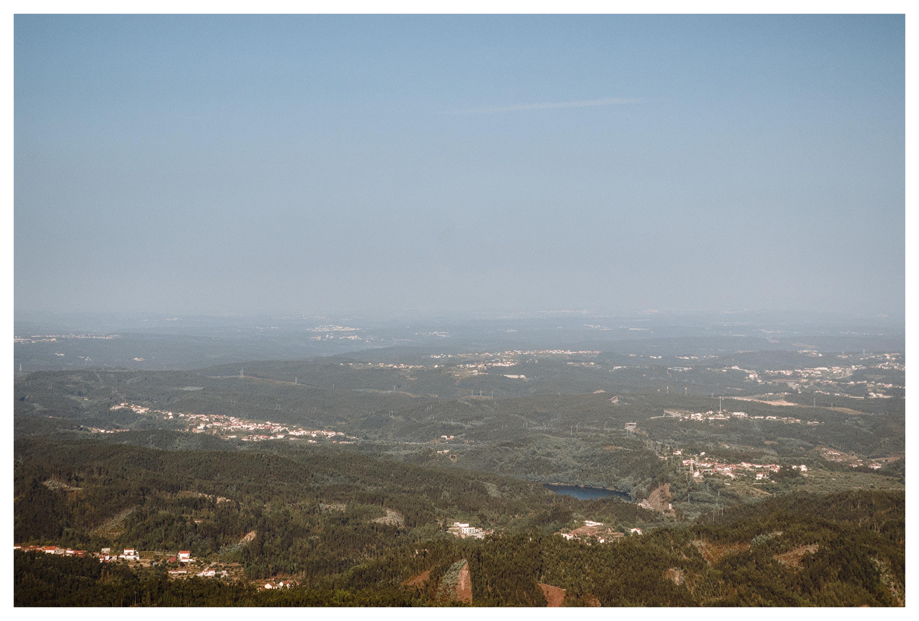 A high-angle, wide view of a vast landscape of rolling green hills and dense forests under a clear, light blue sky. Scattered throughout the valleys and hillsides are several small clusters of white and light-colored light-colored buildings. A small blue body of water is visible nestled within a valley. The landscape stretches toward a hazy horizon where the distant land meets the sky.