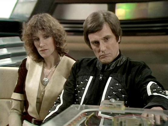 ChatGPT4 describes the image as: "The image features two individuals seated in what appears to be a futuristic or sci-fi setting, likely a spaceship or control room. Both are in costumes that suggest they are characters from a science fiction series. The woman is wearing a light-colored outfit with dark trim, while the man is in a black suit with metallic and white accents. The surrounding environment includes technical equipment and a sleek design typical of a space or high-tech genre. The moo…