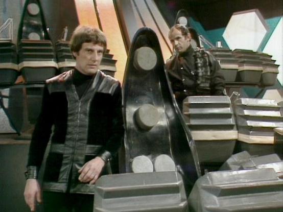 ChatGPT4 describes the image as: "The image shows a scene from a science fiction setting. Two individuals are visible, both dressed in futuristic, dark-colored costumes with metallic or leather-like elements. They are surrounded by what appear to be large, geometric shapes or props, suggesting a spaceship or advanced technological environment. The backdrop includes angular designs and lighting that enhances the sci-fi atmosphere. The context seems to be from a television show or movie, evoking …