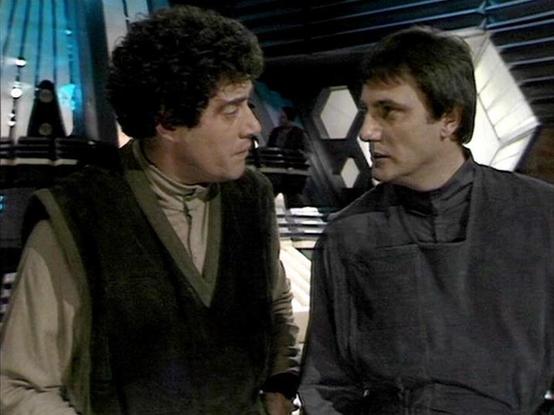 ChatGPT4 describes the image as: "This image is a scene from the British sci-fi television series "Blake's 7." The setting appears to be the interior of a spaceship, with futuristic design elements such as hexagonal patterns and advanced control panels in the background. The two individuals are wearing space-themed costumes, typical of a science fiction genre from that era. The context suggests they are having a serious conversation, possibly discussing a mission or plan aboard the ship."

Bl…