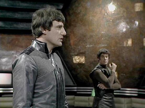 ChatGPT4 describes the image as: "The image shows two individuals in a futuristic setting, likely from a science fiction TV show or movie. The setting appears to be aboard a spaceship, with sleek, metallic walls and a large, spherical, textured background. The individuals are wearing costumes that include leather or synthetic materials, suggesting a sci-fi context. The style and design elements hint at a production from the late 70s or 80s. Without being able to identify specific individuals, I…