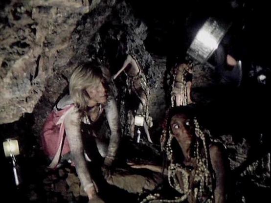 ChatGPT4 describes the image as: "The image appears to be from a scene set in a cave. The setting is dark and rocky, illuminated by a few lights suggesting an underground or mysterious environment. Two characters are visible, one with light hair wearing a pink outfit and another with darker features adorned with intricate, possibly alien-like designs. This scene likely involves an adventurous or possibly sci-fi context, where the characters are exploring or interacting within a subterranean env…