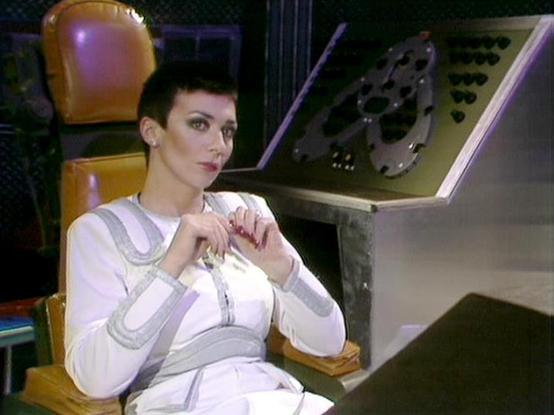 ChatGPT4 describes the image as: "The image depicts a person sitting in what appears to be a futuristic or sci-fi setting. The individual is wearing a white outfit with silver accents, which suggests a space or sci-fi theme. Behind them is a control panel filled with knobs and buttons, enhancing the high-tech environment. The seat and surrounding decor have a retro-futuristic design, commonly seen in science fiction shows from past decades. The overall atmosphere is one of command and technolog…