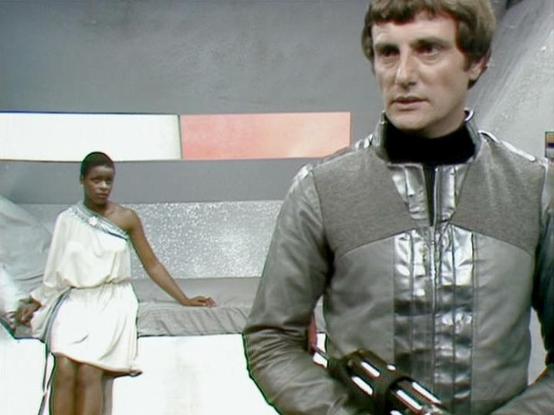 ChatGPT4 describes the image as: "The image features two individuals in a futuristic setting reminiscent of a sci-fi TV show. The person in the foreground is wearing a metallic, space-themed outfit and holding a futuristic weapon, suggesting a tense or serious situation. The background shows another individual, wearing an elegant white outfit with decorative elements, seated on a modernistic, metallic-looking structure. The setting includes geometric patterns and metallic hues, typical of a spa…