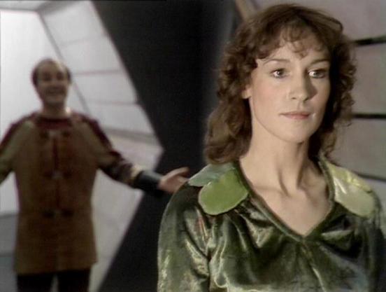 ChatGPT4 describes the image as: "The image appears to depict a scene from a science fiction setting, possibly on a spaceship or futuristic environment. A woman is in the foreground, wearing a metallic-looking green outfit with distinctive shoulders. Behind her, in the background, is a man who seems to be gesturing or speaking, dressed in a tunic-like outfit. The architecture includes angled walls, contributing to the futuristic ambiance.

Michael Keating and Jan Chappell were actors in the Bri…