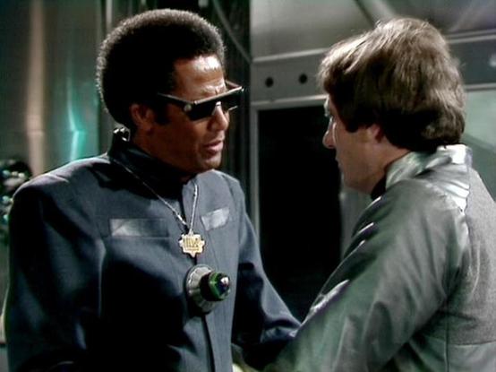 ChatGPT4 describes the image as: "The image appears to be from a science fiction television setting. It shows two individuals in conversation, dressed in futuristic attire with distinctive accessories. The man on the left is wearing sunglasses and has a stylish badge around his neck. The setting suggests a high-tech or space-themed environment, possibly on a spaceship or a futuristic facility, given the metallic and industrial backdrop. The clothing and props suggest a show from the late 20th c…