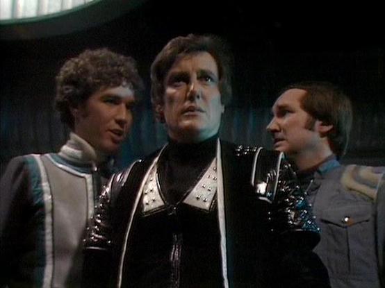 ChatGPT4 describes the image as: "The image features three men in a futuristic setting, which appears to be a science fiction environment, possibly from a television show. The setting has a dimly lit interior with metallic and sleek design elements, suggesting a spaceship or high-tech facility. The men are dressed in distinct, futuristic costumes, likely indicating their roles as characters in a sci-fi series. The central figure wears a black costume with metallic and shiny details, while the o…