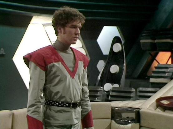 ChatGPT4 describes the image as: "The image depicts a person standing in a science fiction setting. The individual is wearing a futuristic outfit with red and gray colors, featuring a V-shaped design on the chest and shoulders, and a belt. The background suggests a spaceship or high-tech environment with geometric structures and panels. The lighting is dim, consistent with the interior of a spacecraft or control room. The overall atmosphere is typical of a classic sci-fi television series."

…