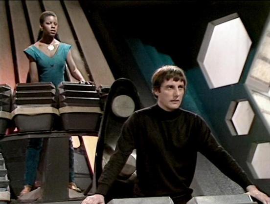 ChatGPT4 describes the image as: "The image shows a scene from a science fiction television show set. It appears to be inside a futuristic spacecraft, characterized by angular and geometric patterns on the walls and high-tech controls. There are two individuals: one standing at what looks like a control panel, wearing a teal outfit, and another seated closer to the viewer, wearing a black top. The overall atmosphere suggests a dramatic or intense moment, typical of many sci-fi narratives."

B…