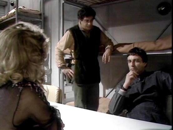 Claude 3.7 describes the image as: "This image appears to be from a science fiction television series, set in what looks like a spacecraft or space station interior. The scene takes place in what appears to be crew quarters with bunk beds visible in the background.

The image shows three people in conversation. One person is standing in the center wearing a dark vest over a light-colored shirt. To the right, someone is seated in dark clothing with a thoughtful expression. On the left, partially…