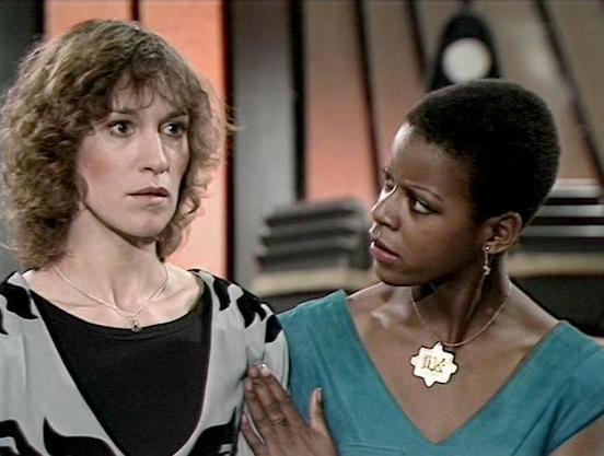 Claude 3.7 describes the image as: "The image shows a scene from what appears to be a vintage science fiction television production from the late 1970s or early 1980s. Two people are engaged in what seems to be a serious conversation. On the left is a person with curly hair wearing a black and white patterned outfit over a dark top. On the right is a person with short hair wearing a teal/turquoise top and a distinctive star-shaped pendant.

The setting appears to be some kind of futuristic or s…