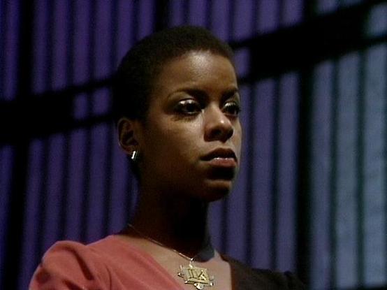 Claude 3.7 describes the image as: "This image shows a portrait of a person with short dark hair wearing a pink/salmon-colored top and a distinctive star-shaped pendant necklace. The lighting creates a dramatic effect with purple-tinted vertical bars or blinds visible in the background, giving the scene a science fiction atmosphere. 

The composition features a close-up shot with the subject gazing slightly off-camera with an intense, serious expression. The cinematography has that characterist…