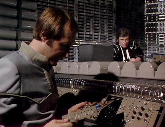 Claude Sonnet 4.0 describes the image as: "This image shows a scene set in what appears to be the flight deck or control room of a spacecraft. The setting features a futuristic control console with various buttons, switches, and monitoring equipment typical of science fiction television production design from the late 1970s. Two crew members are visible - one in the foreground operating controls at the main console, wearing what appears to be a light-colored uniform, while another crew member c…