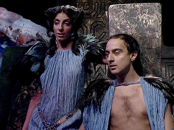 Claude 3.7 describes the image as: "This image appears to be from a science fiction television production, showing two performers in elaborate costume design. They're wearing distinctive blue-gray fringed garments with dark feathered or fur-like elements at the shoulders. The costumes have a ceremonial or ritualistic quality, with one outfit featuring an open chest design and the other a more covered style. The setting features metallic wall panels with geometric patterns in the background, sug…