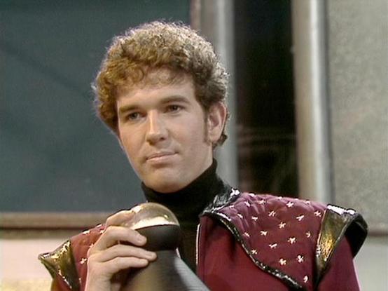 Claude Sonnet 4.0 describes the image as: "I can see this is an image from a science fiction television series showing someone in what appears to be a futuristic military or space-faring uniform. The costume features a distinctive burgundy or maroon colored tunic with decorative metallic studs or embellishments, and black accents at the collar. The person has curly brown hair and is holding what looks like a futuristic weapon or device. The setting appears to be aboard a spacecraft or space sta…