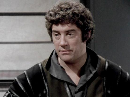 Claude 3.7 describes the image as: "The image shows a person with curly dark hair wearing what appears to be a dark costume with distinctive panels or stripes. The styling and production quality suggests this is from a classic science fiction television production, likely from the late 1970s or early 1980s based on the visual aesthetic.

The setting appears to be some kind of interior space, possibly a spacecraft or futuristic facility, with a simple, somewhat utilitarian background typical of …